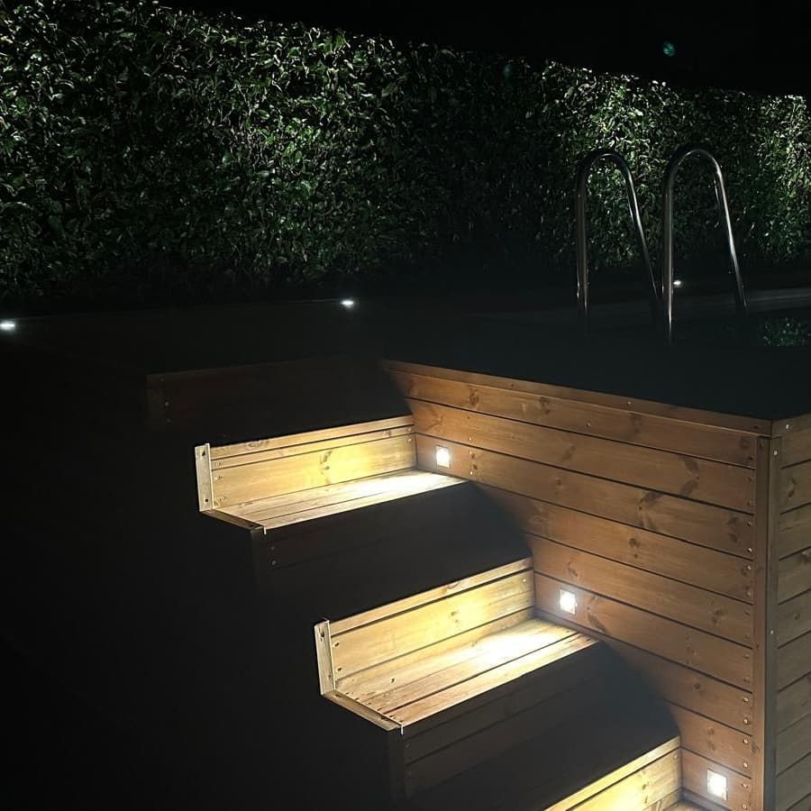 Moderna scalinata esterna in legno con calda illuminazione a LED notturna, accanto a una siepe e a una piscina.