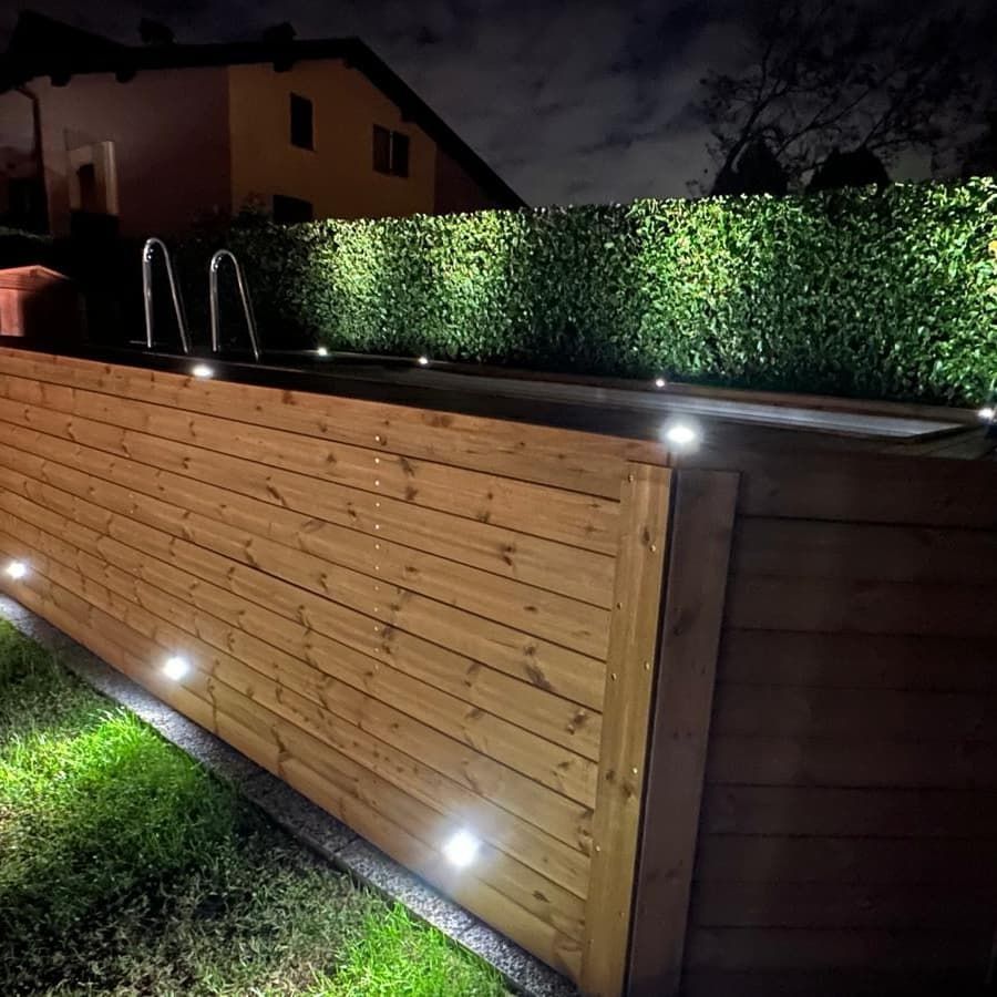 Vista notturna di un moderno cortile con recinzione in legno, siepe e luci a terra che illuminano il prato.