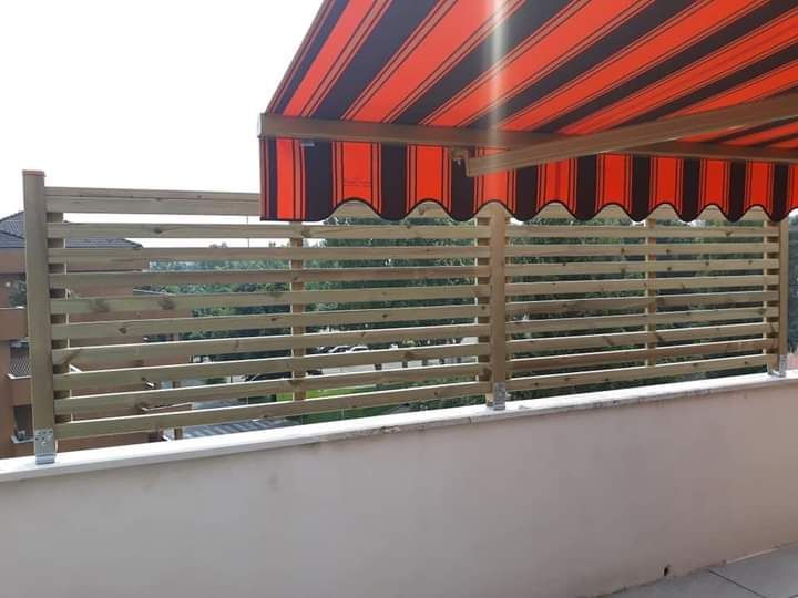 Balcone con ringhiera orizzontale grigia e tenda da sole a righe rosse e nere sopra una parete bianca.