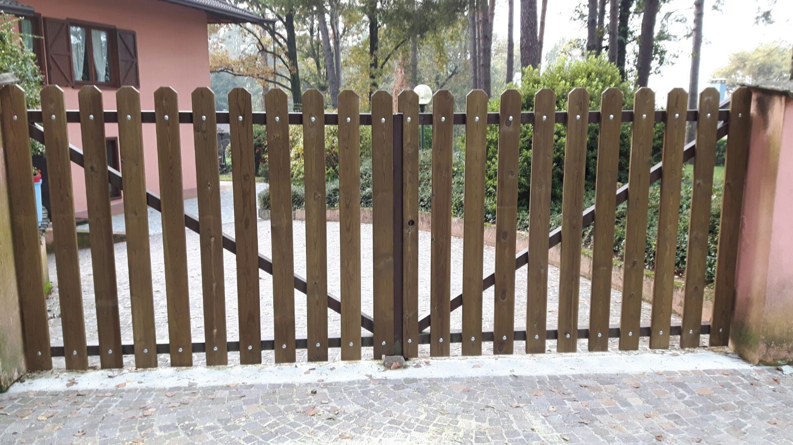 Cancello in legno a paletti che attraversa un vialetto accanto a una casa e ad alberi.