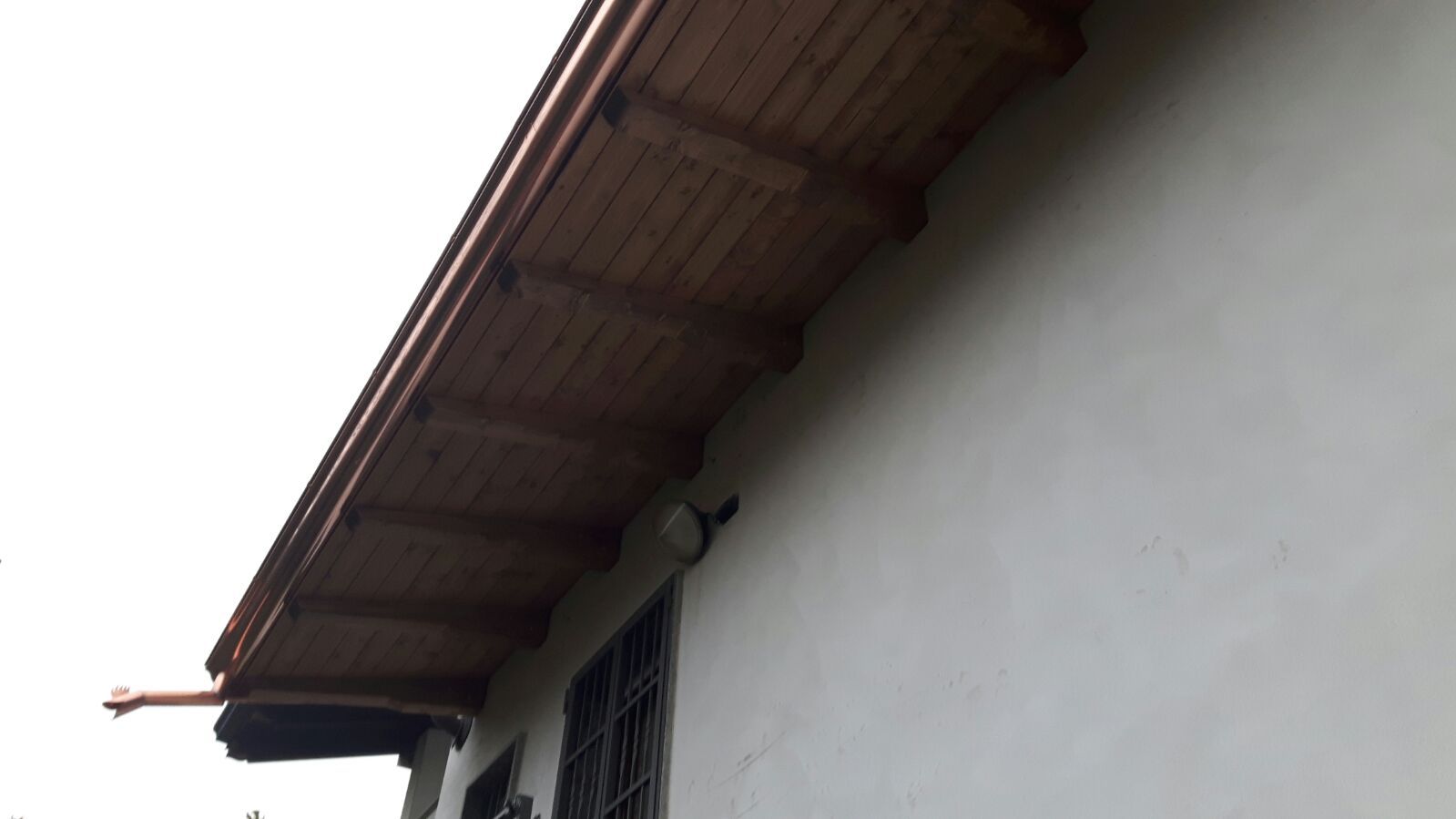 Vista laterale di una parete di una casa bianca con una sporgenza del tetto in legno marrone e inferriate scure alle finestre.