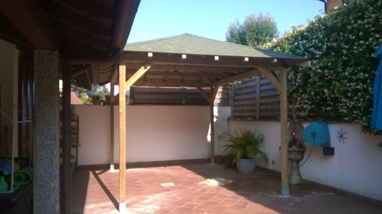 Patio soleggiato con pergolato in legno, pavimento piastrellato, parete bianca e piante in vaso.