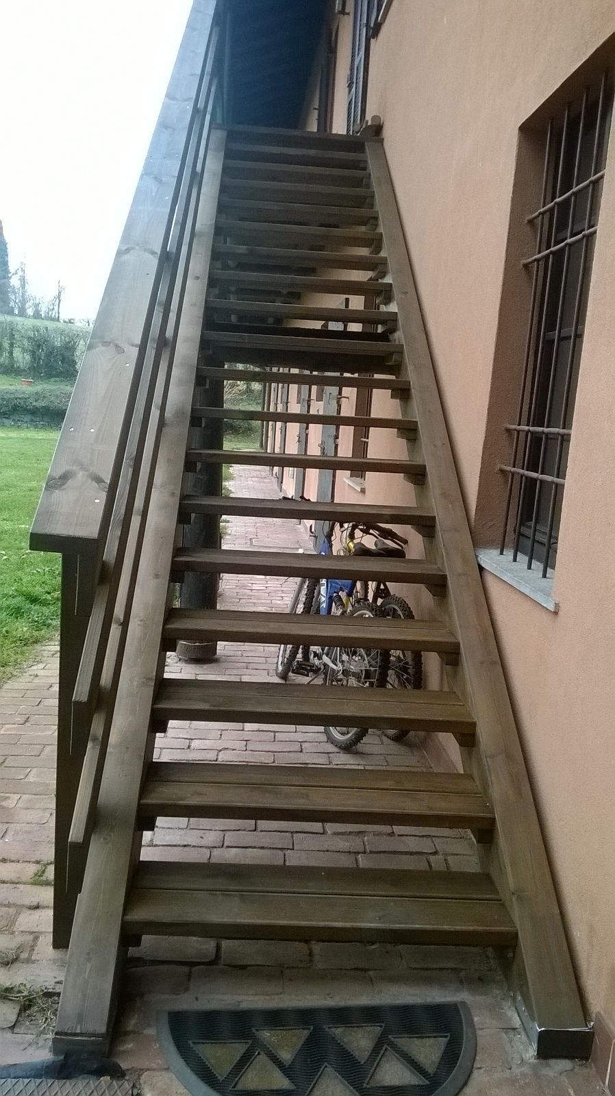 Ripida scala esterna in legno accanto a un edificio beige, con ringhiera in metallo e finestra con sbarre.