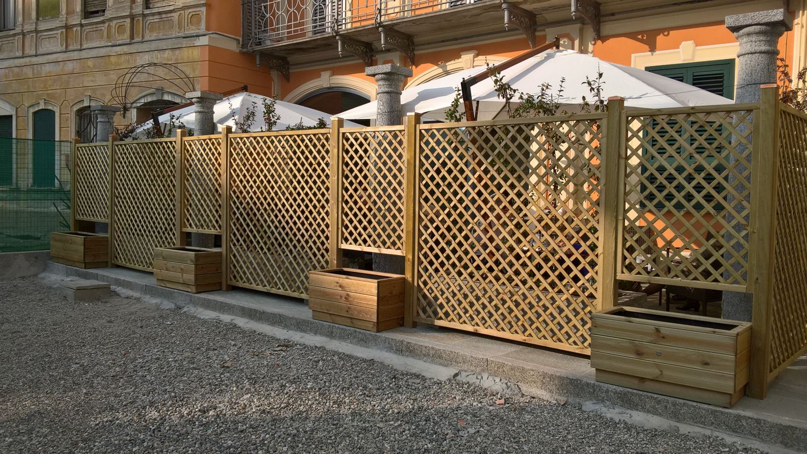 Patio esterno recintato con traliccio e ombrelloni bianchi, adiacente a un edificio in mattoni e a un terreno ghiaioso.