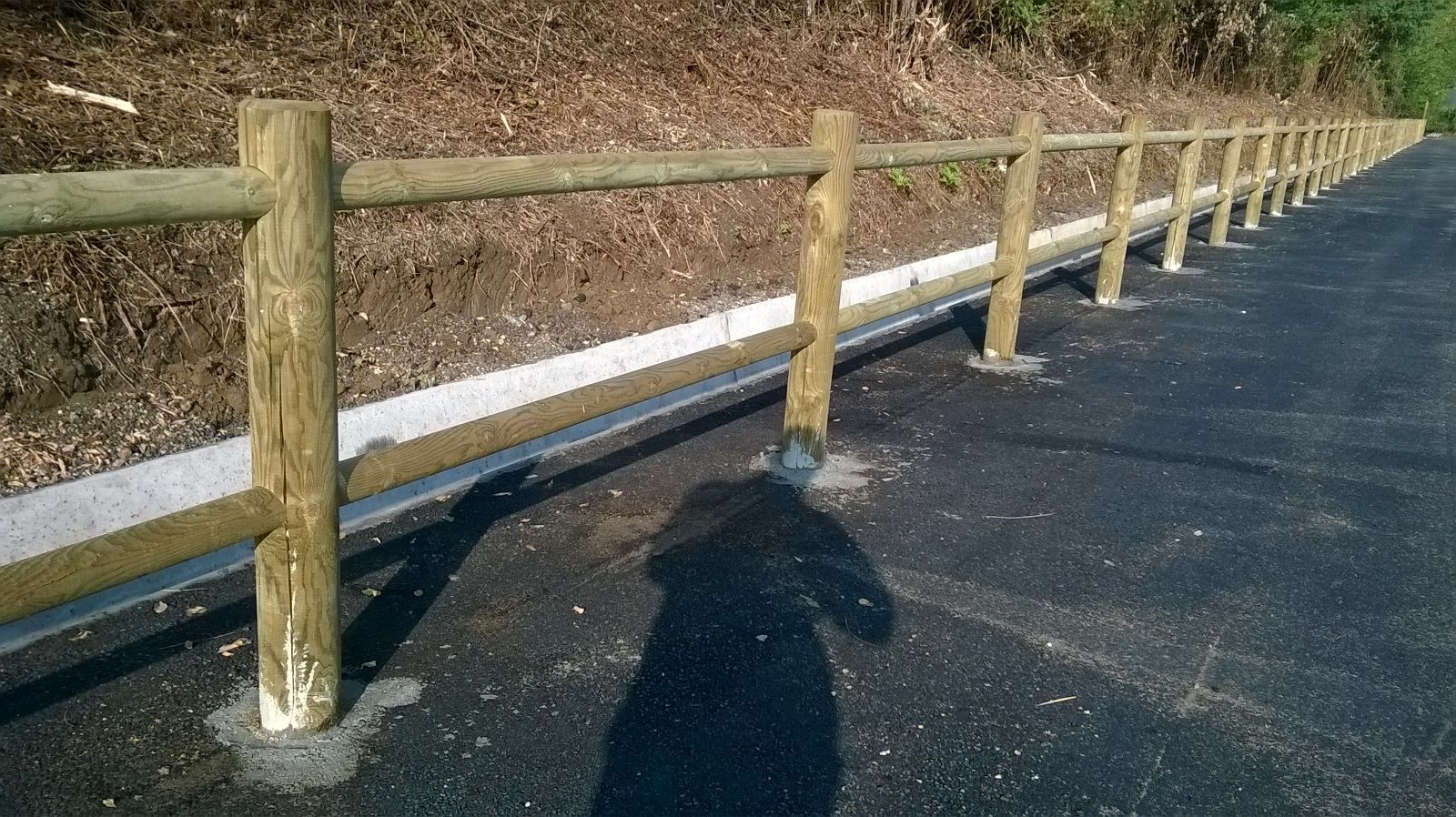 Pali di legno di una recinzione a bordo strada, accanto a un sentiero lastricato, con chiazze di neve e un terrapieno erboso illuminato dal sole.