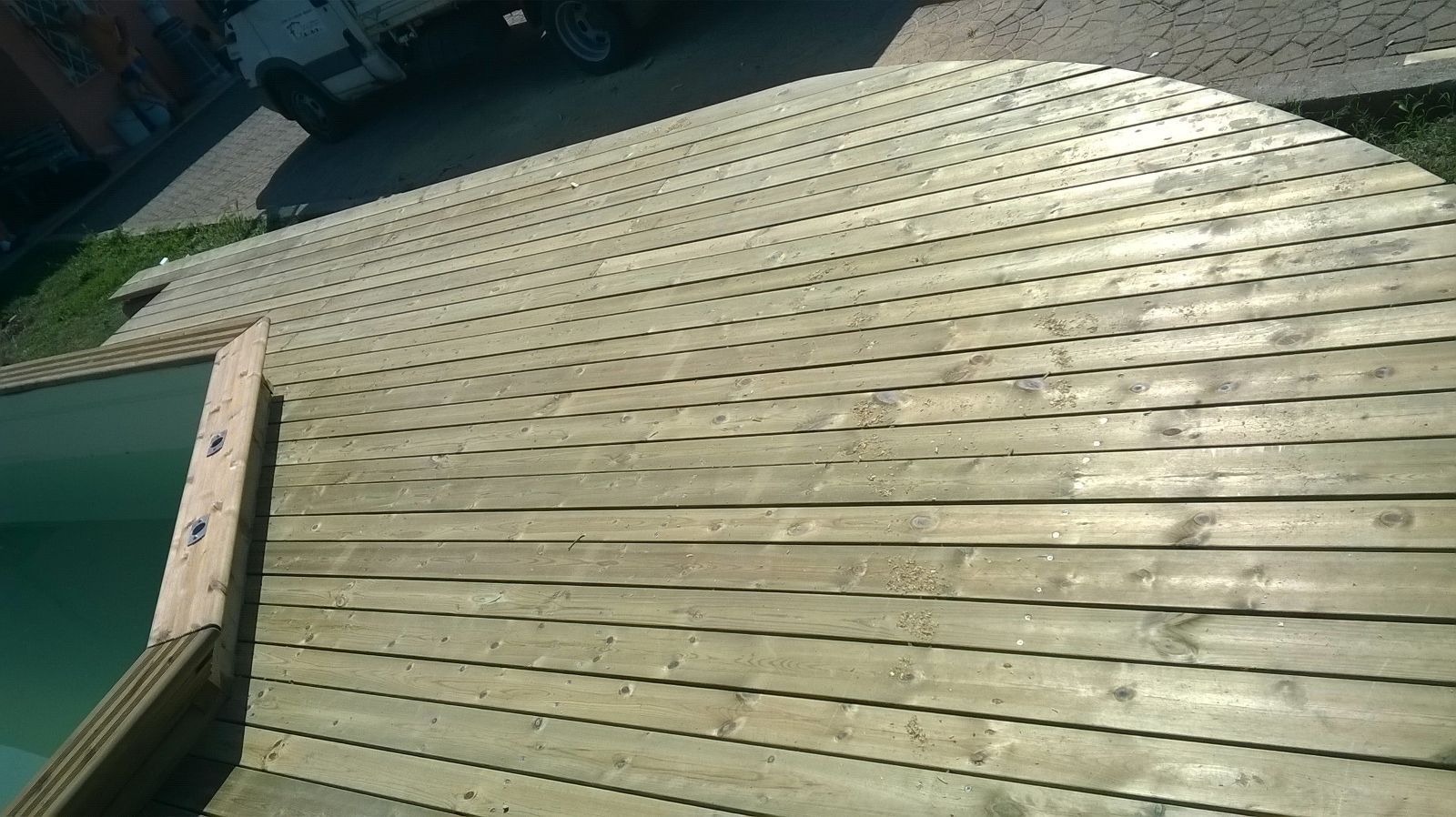 Terrazza in legno curva in costruzione, illuminata da un sole splendente, con assi e apertura della scala ben visibili.