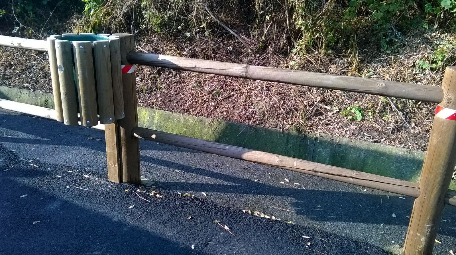Barriera stradale in legno con traversa orizzontale a lato di un sentiero lastricato e di aree verdi.
