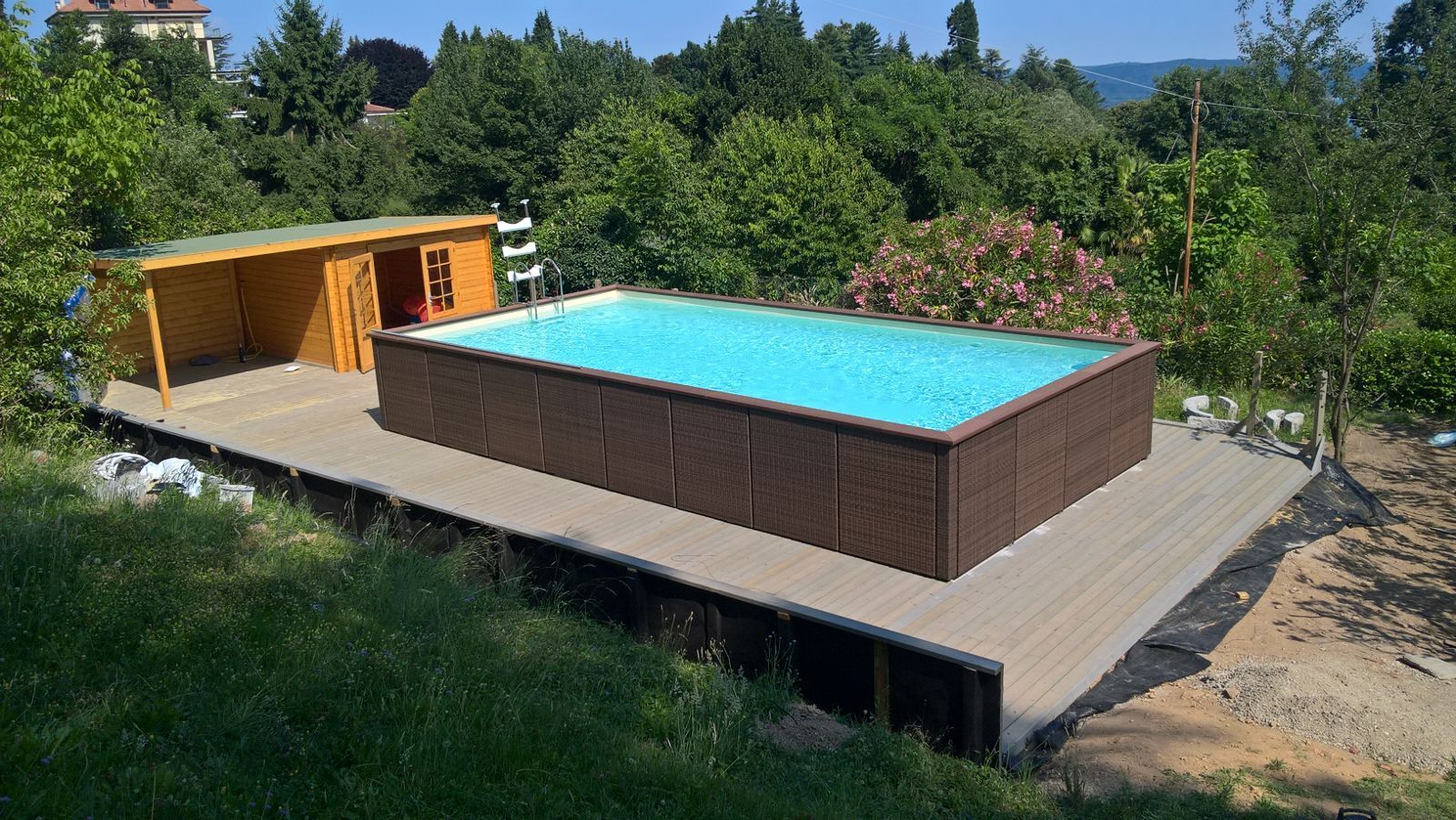 Giardino moderno con piscina fuori terra su una terrazza in legno, circondato da alberi e vegetazione.