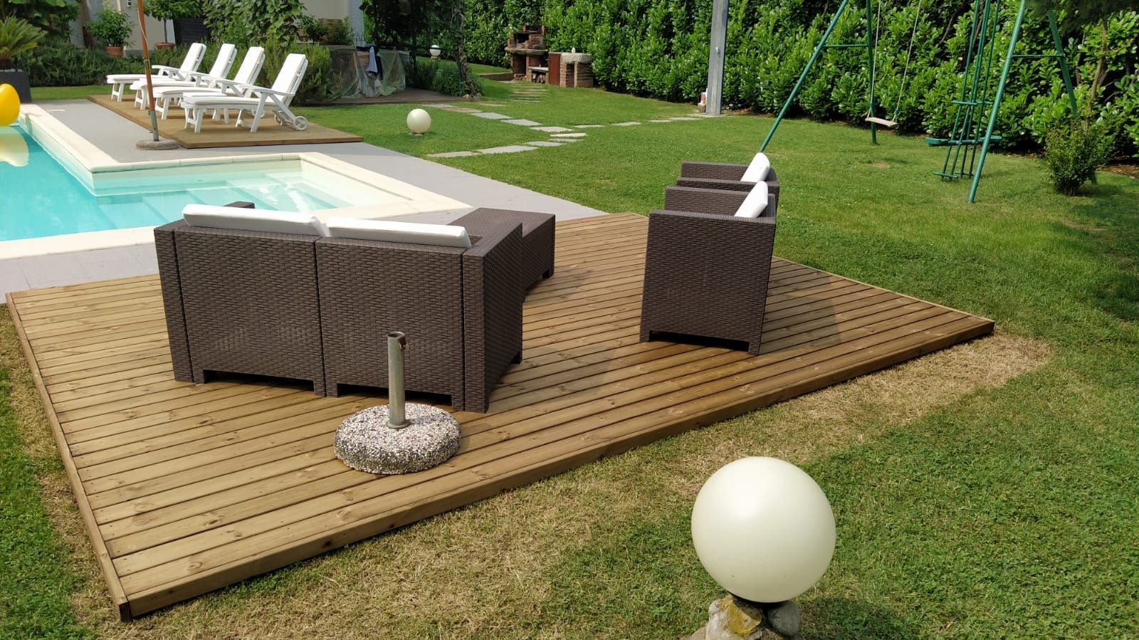Patio esterno con sedie e tavolo in vimini su una terrazza in legno accanto alla piscina e al prato.
