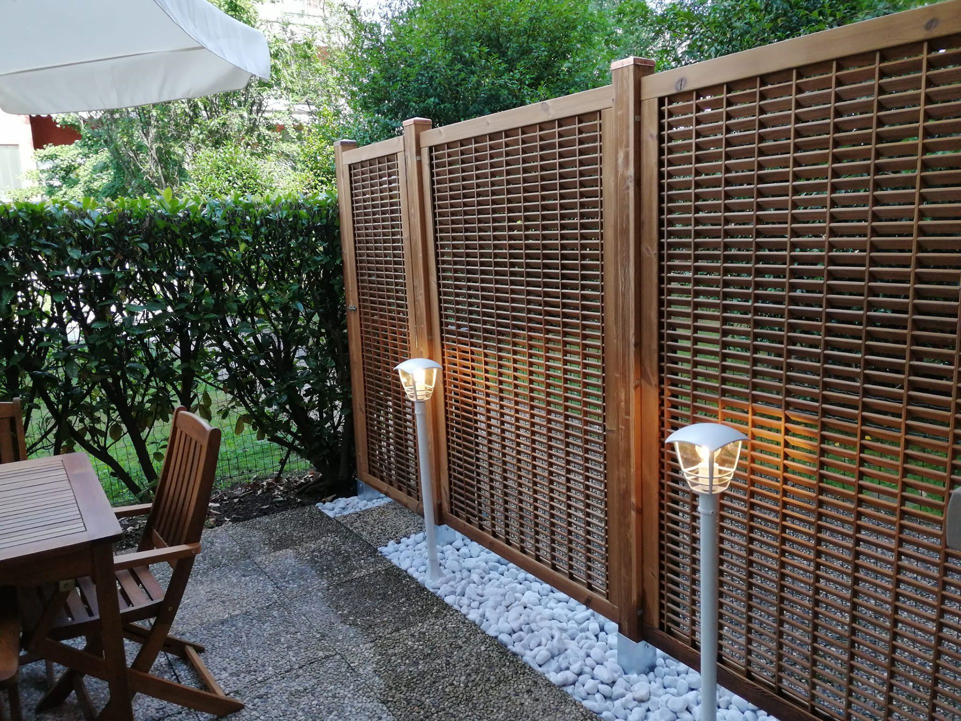 Patio sul retro con recinzione in legno a traliccio, luci esterne, tavolo e sedie su sentiero di ghiaia.