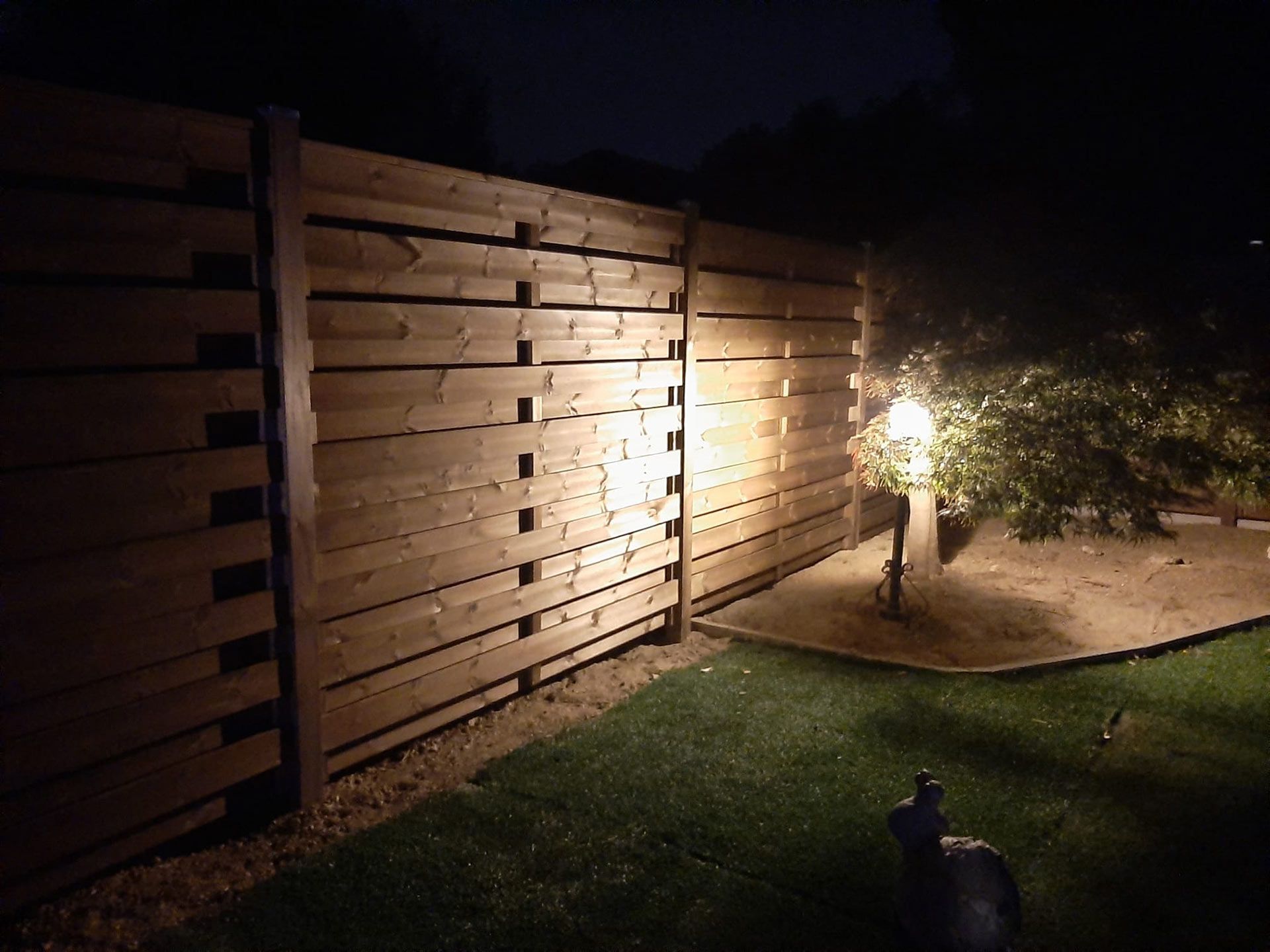 Cortile notturno con recinzione in legno, sentiero illuminato e un cagnolino sull'erba.