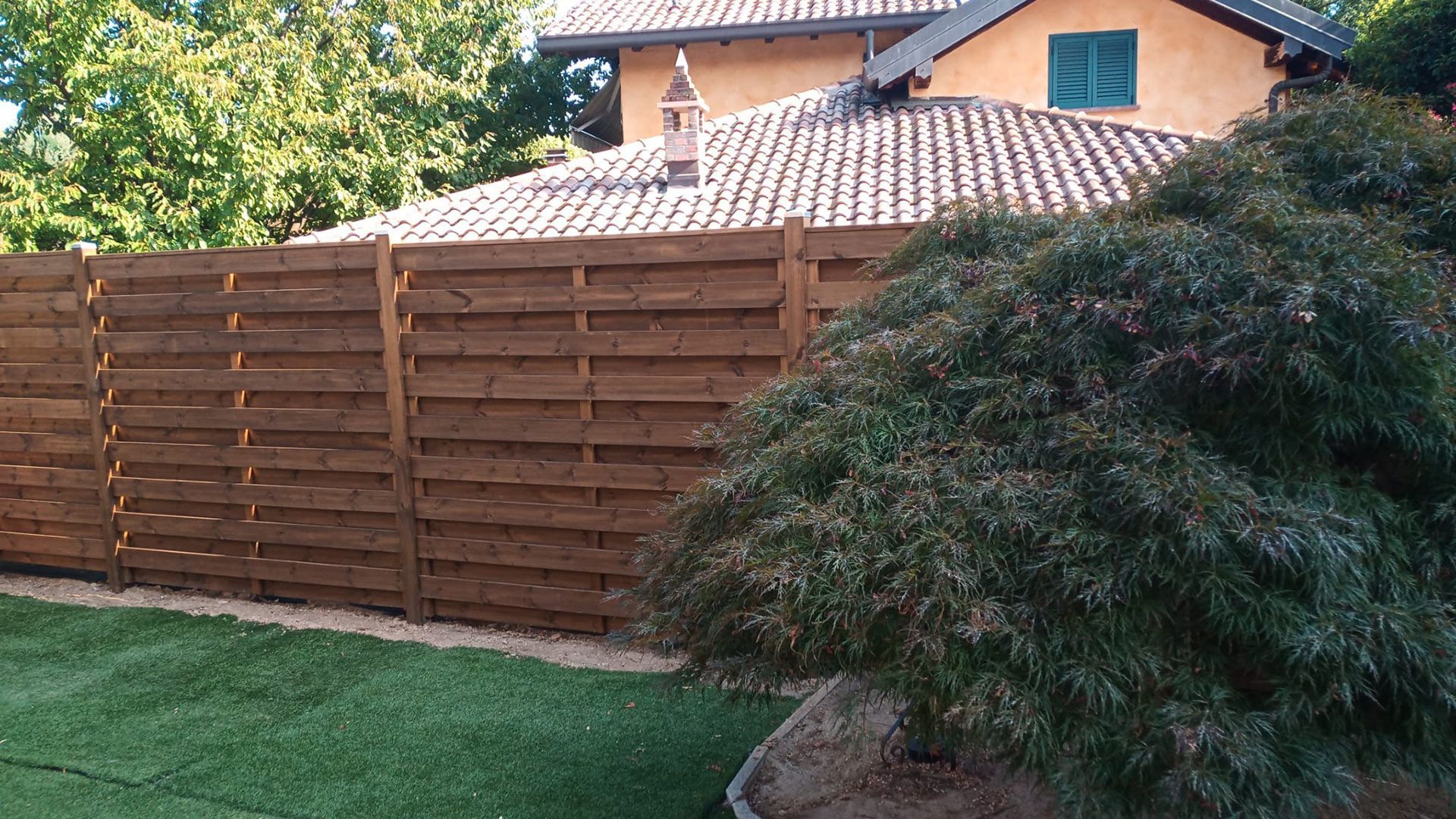 Recinzione in legno per la privacy accanto a un prato verde e a un grande arbusto di fronte a una casa con tetto di tegole.