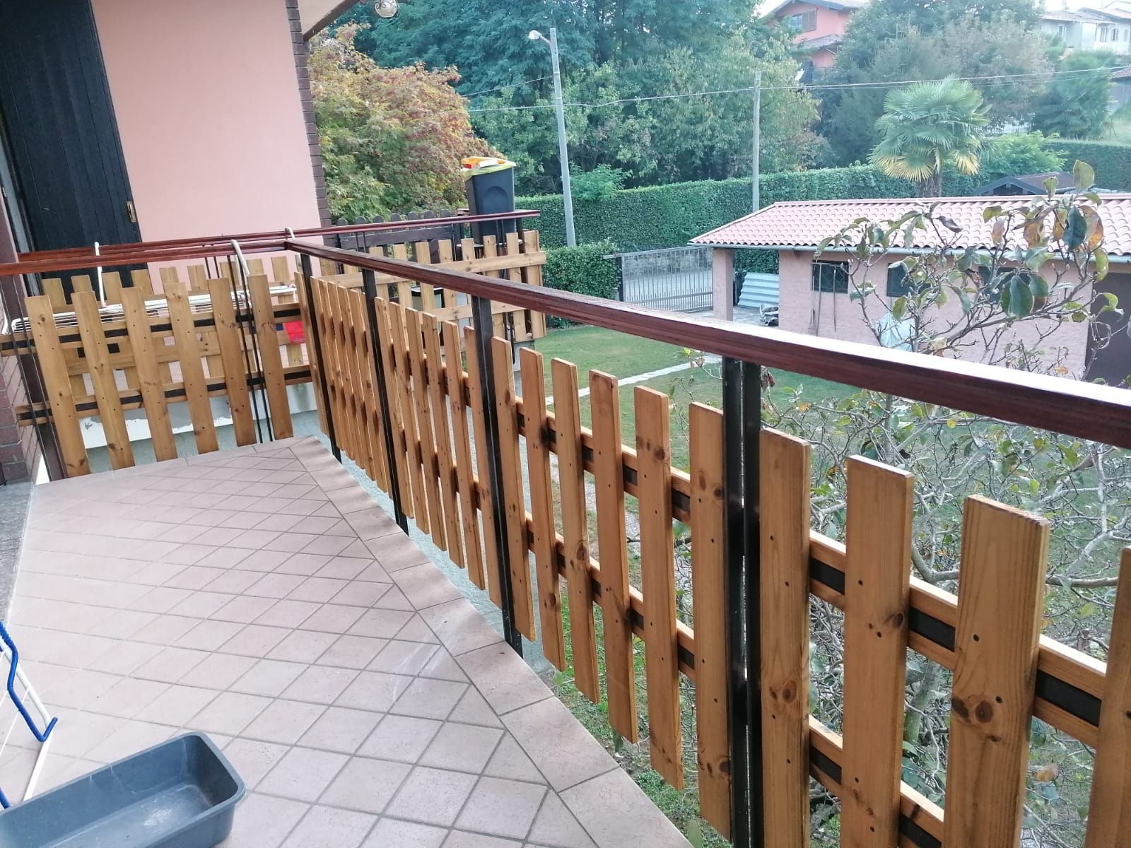 Balcone con pavimento piastrellato e ringhiera in legno con vista sugli alberi e sulle case vicine.