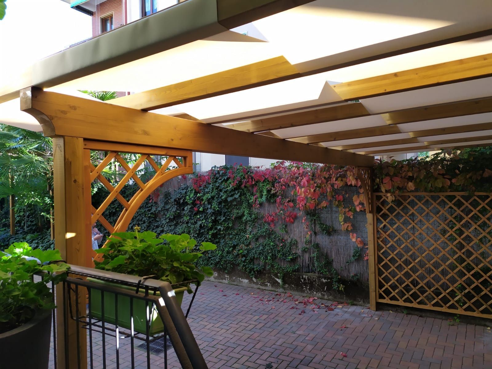 Patio coperto con travi in legno, pergolato e cortile pavimentato in mattoni adiacente a un muro di cinta del giardino.