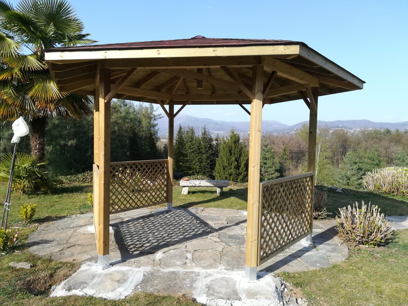 Gazebo in legno con pareti a traliccio su un patio in pietra in un giardino soleggiato con colline sullo sfondo.