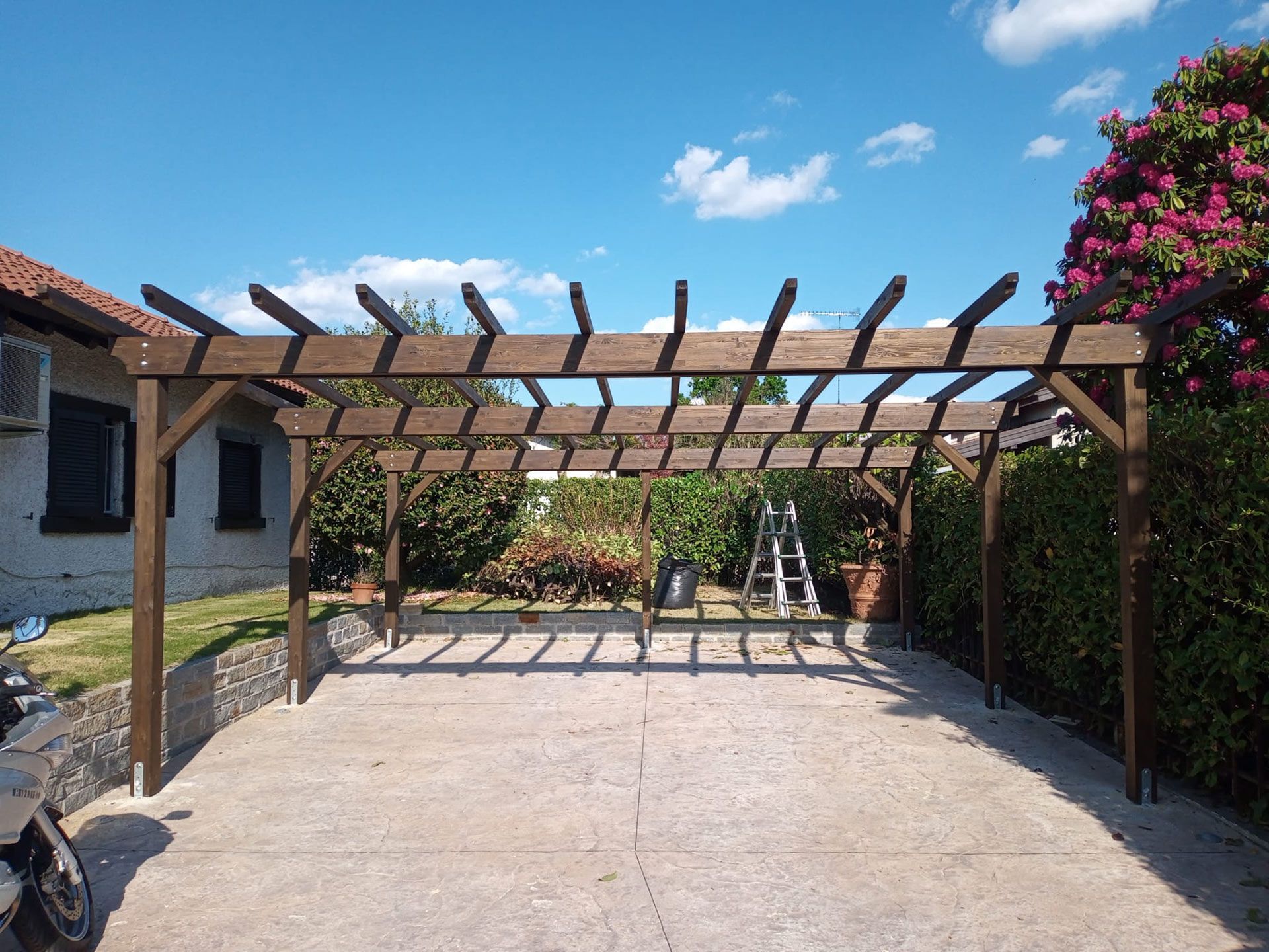 Pergola in legno sopra un vialetto pavimentato accanto a una casa, con cielo azzurro e piante rampicanti in fiore.