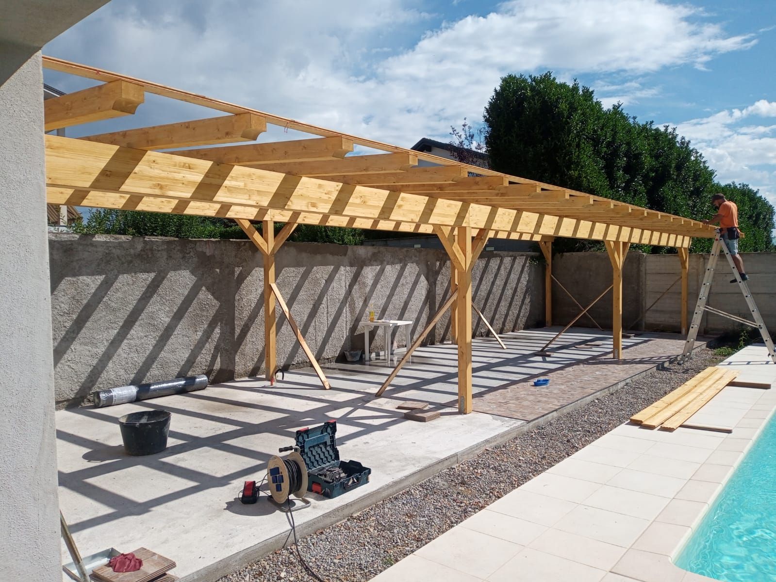 Pergola in legno in costruzione accanto a una piscina, con attrezzi, ghiaia e operai su un patio soleggiato.