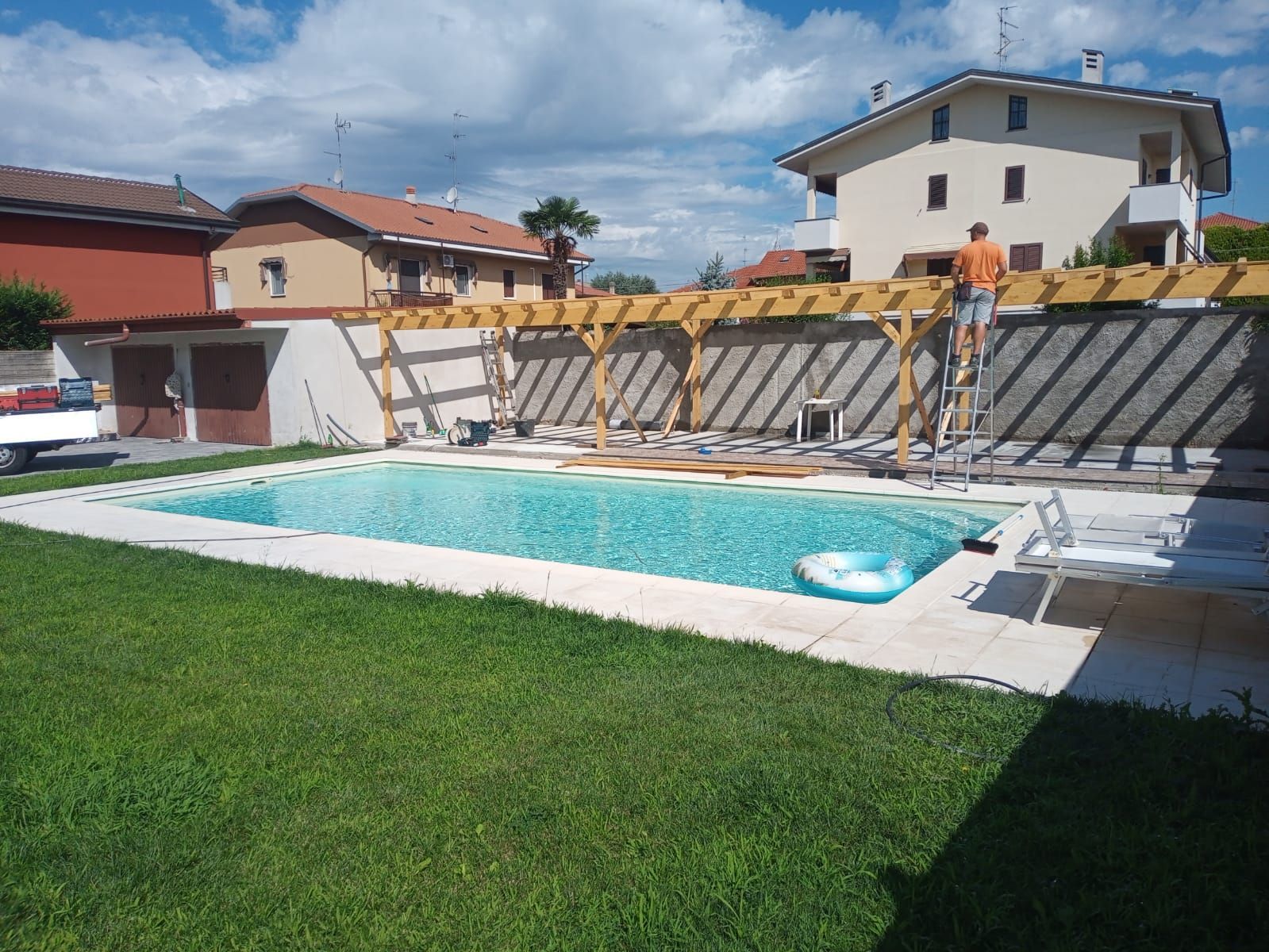 Piscina nel cortile sul retro, accanto a un prato verde, con case e lettini prendisole sullo sfondo.