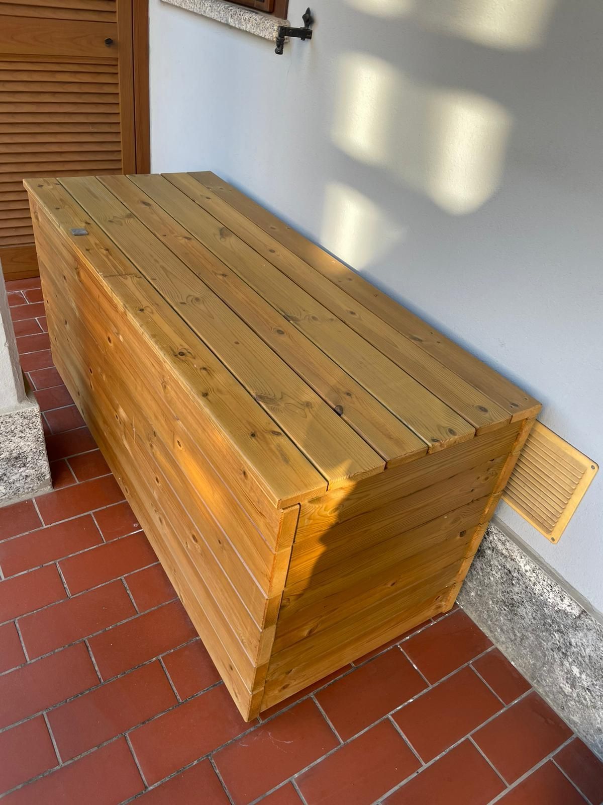 Panca o cassapanca in legno addossata a un muro su un patio piastrellato, esposta al sole.