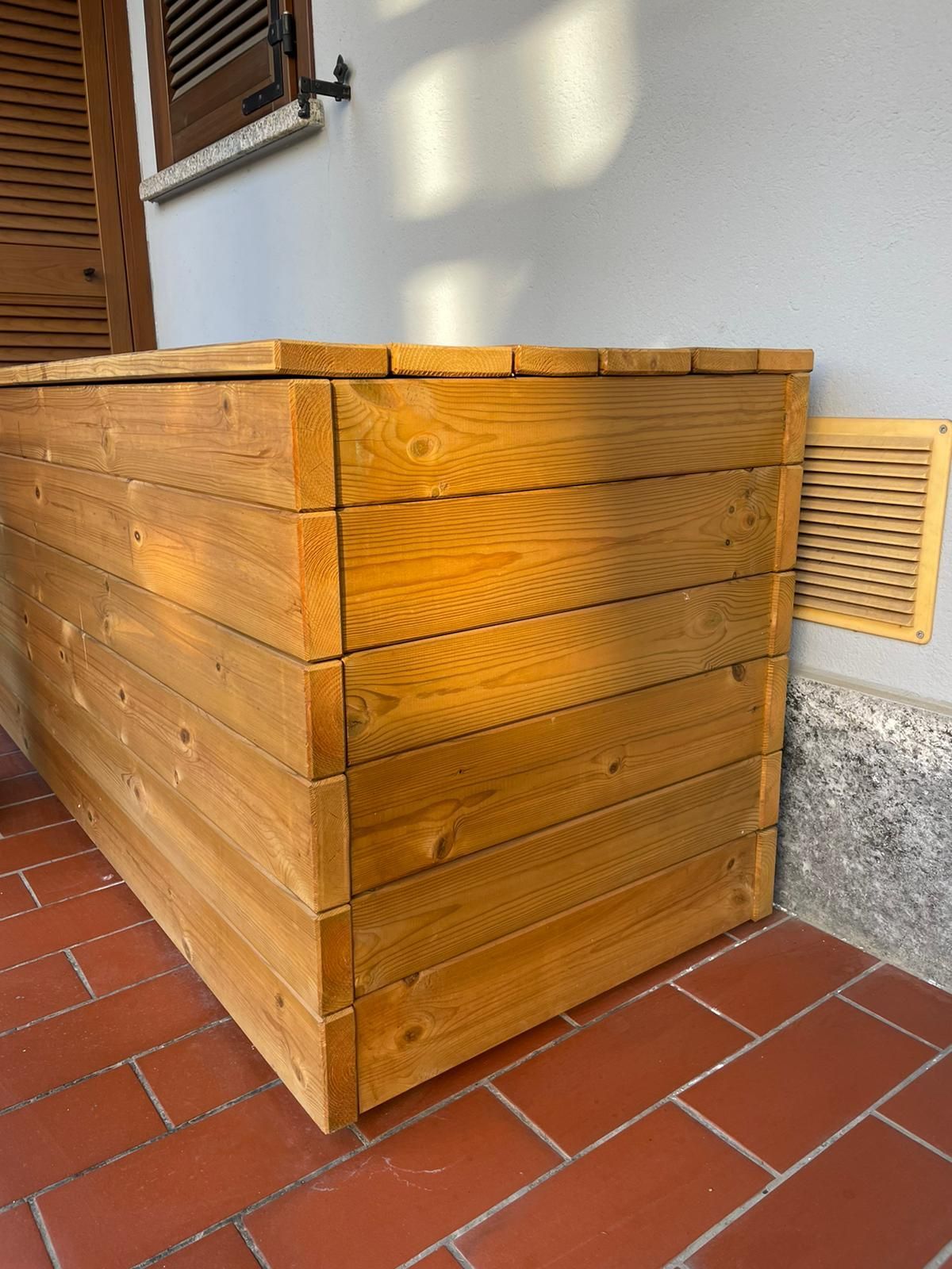 Panca contenitore in legno per esterni, posizionata accanto al muro di un edificio su un patio piastrellato.