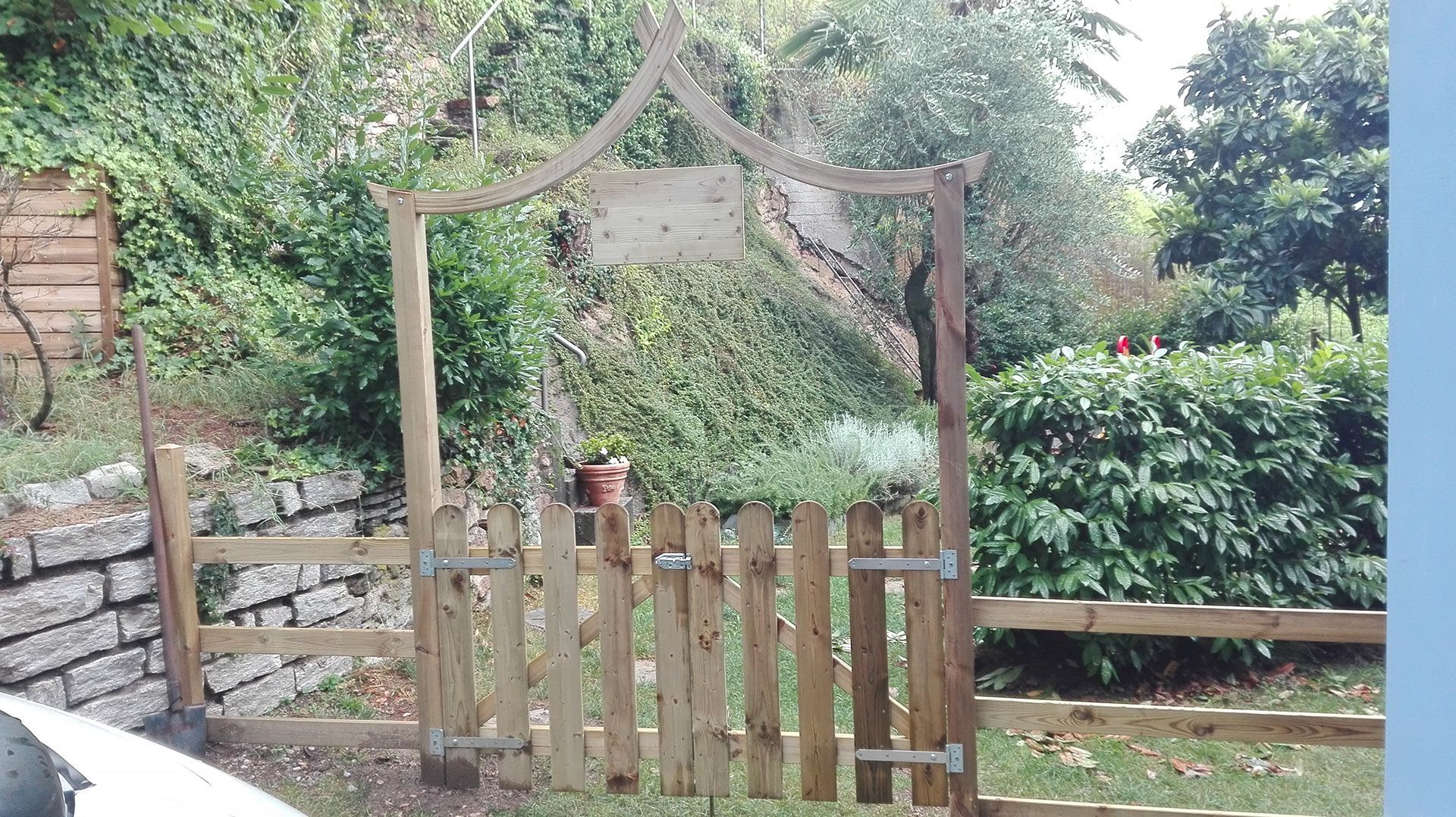 Cancello da giardino in legno e pergolato con vegetazione rigogliosa accanto a un muro di pietra e una siepe.