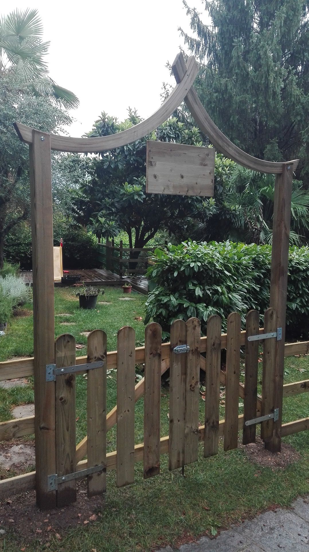 Un rustico cancello in legno che conduce a un rigoglioso giardino, incorniciato da un arco decorativo e da un cartello appeso.