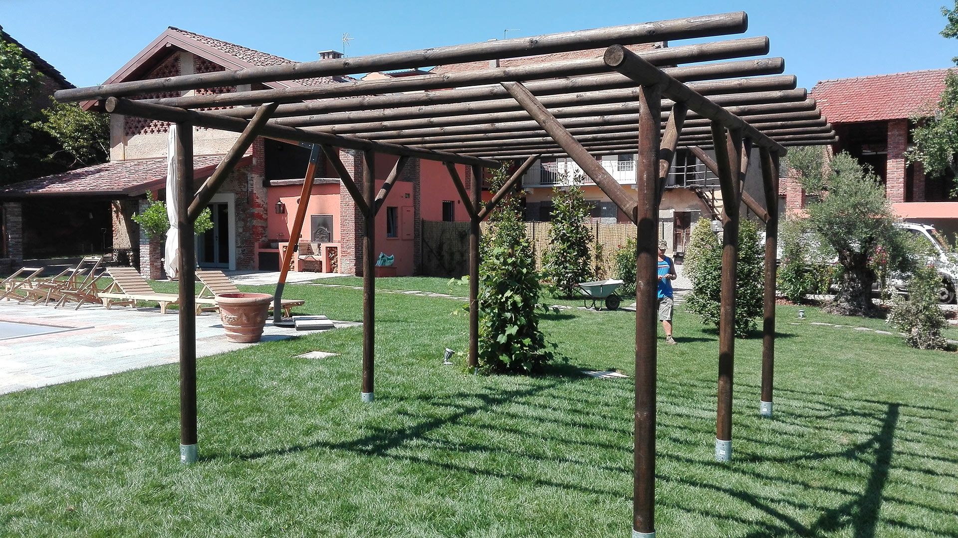 Pergola in legno in un cortile erboso con edifici dai tetti rossi sullo sfondo.