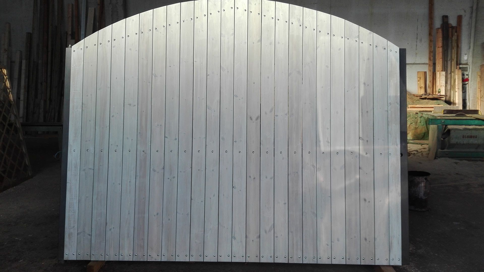 Muro o cancello in legno ad arco bianco in un cortile industriale poco illuminato, con tubi e attrezzature sullo sfondo.