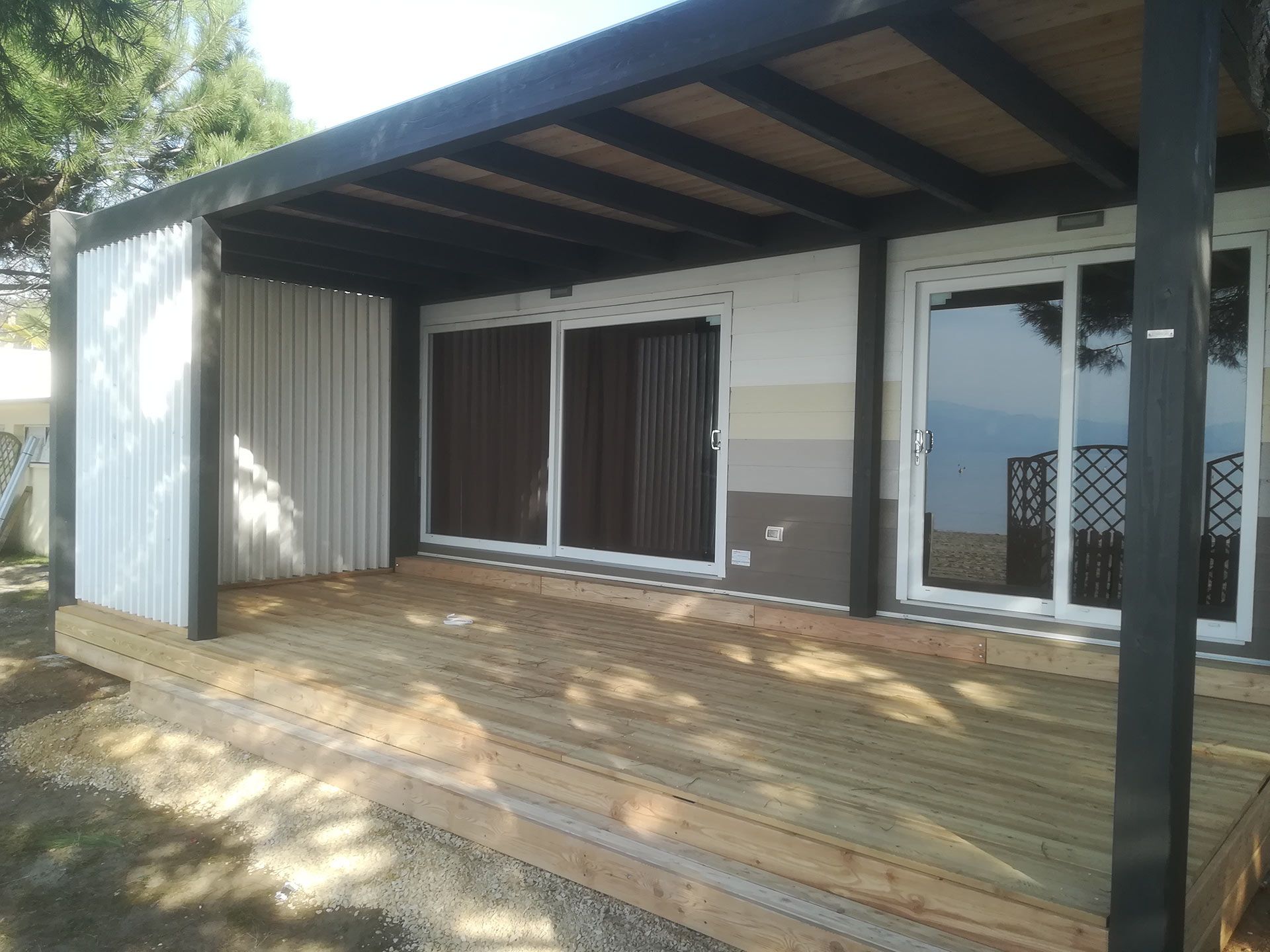 Veranda di una piccola casa con terrazza in legno, tetto scuro, rivestimento esterno bianco e porte scorrevoli in vetro.