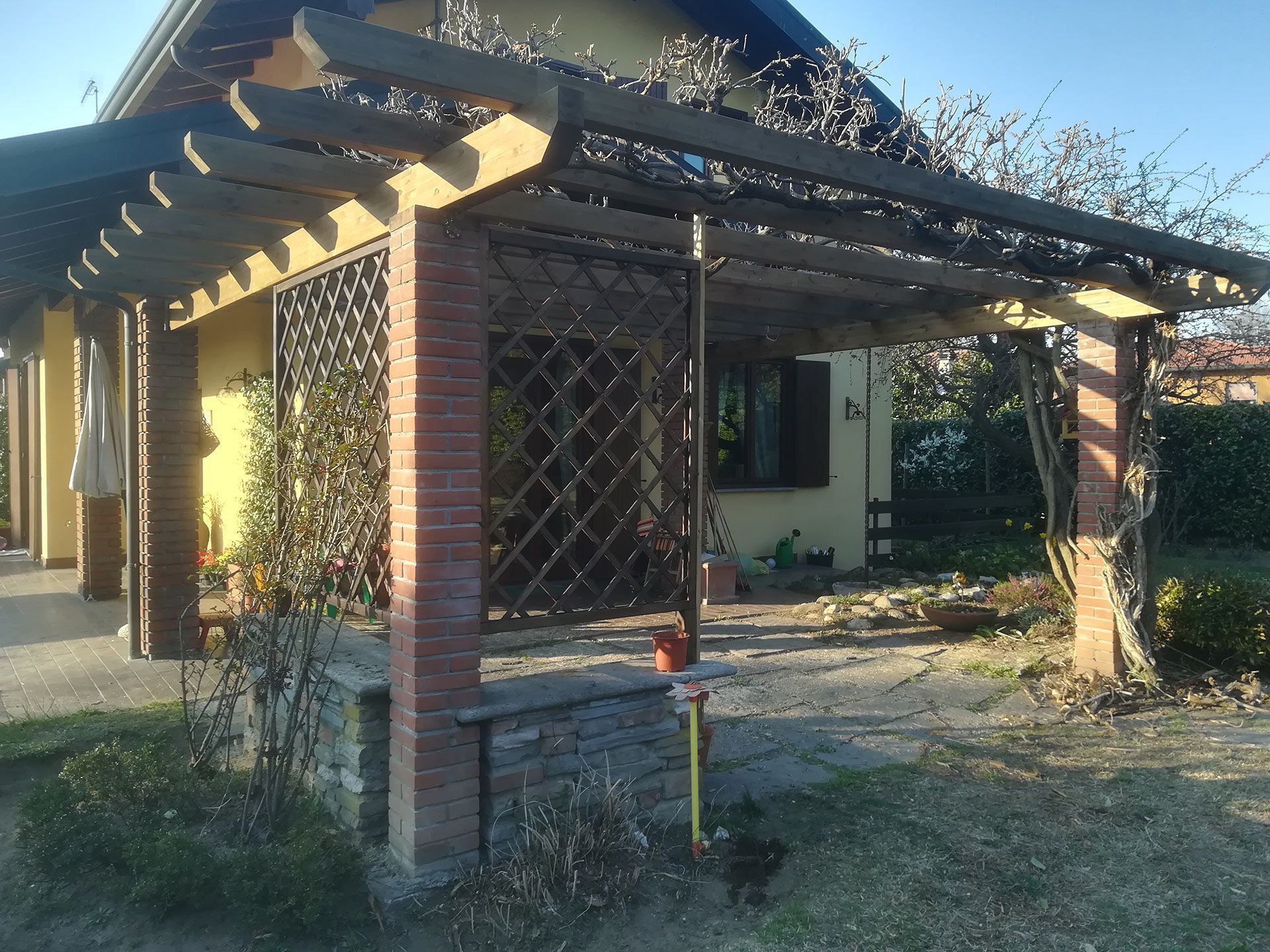 Ingresso rustico di una casa con pergola in legno, pilastri in mattoni e piante rampicanti in un cortile soleggiato.