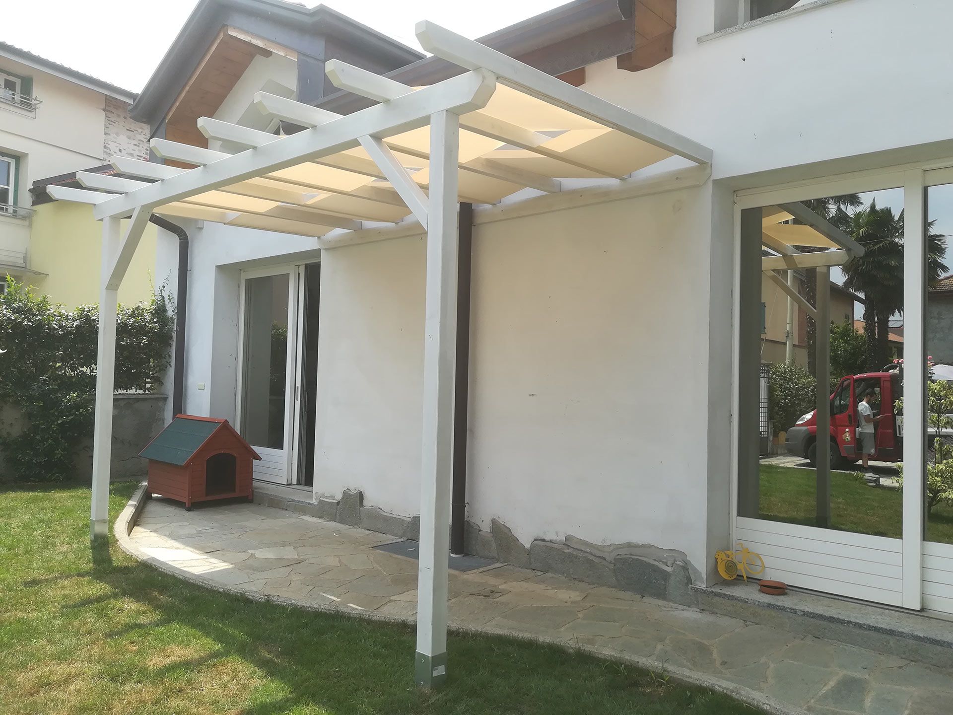 Patio sul retro con pergola bianca, porte scorrevoli in vetro e una piccola cuccia rossa addossata a una parete della casa.