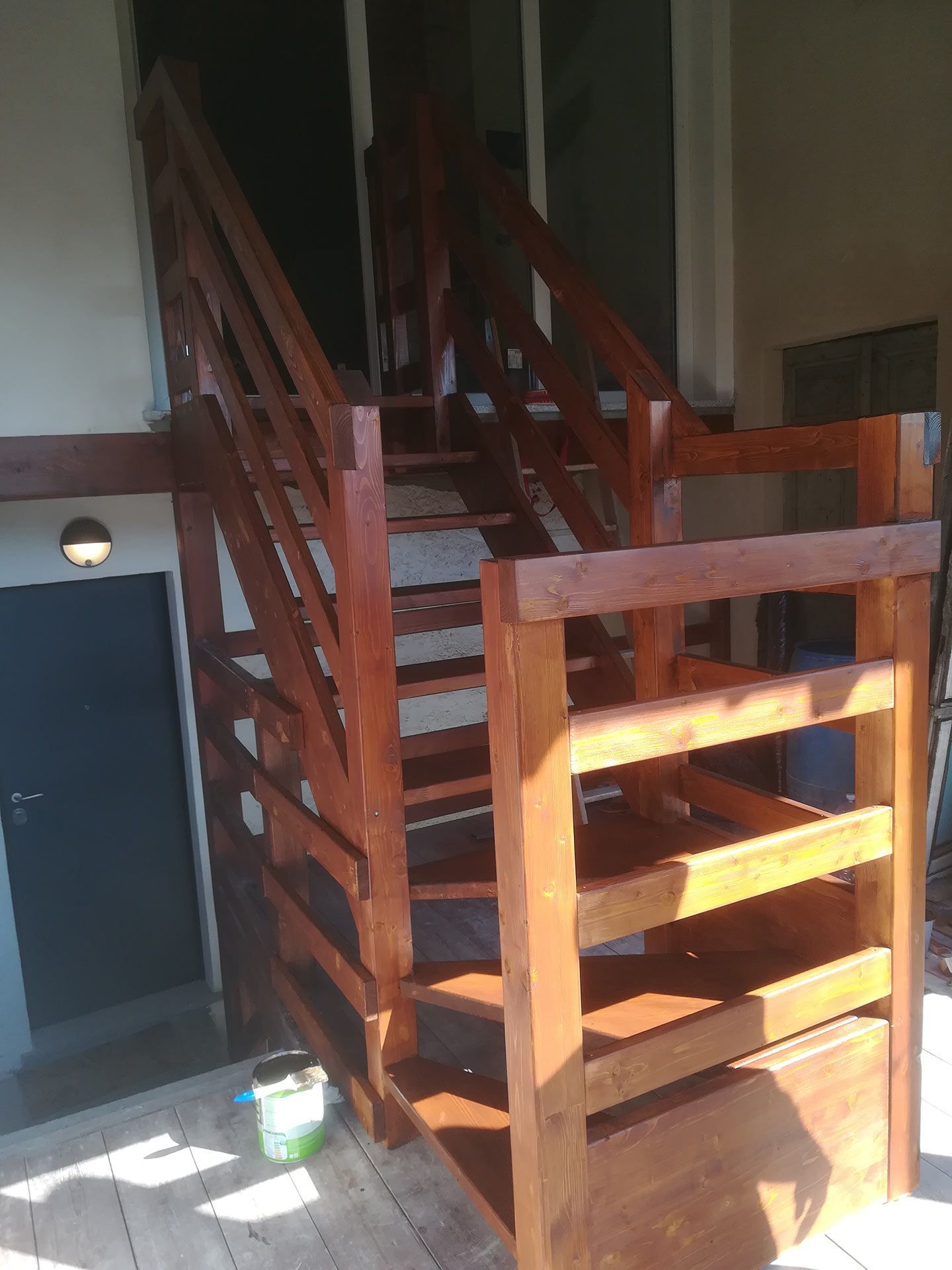 Scala in legno con ringhiera rossa in un interno poco illuminato, rischiarato dalla luce del sole proveniente da sinistra.