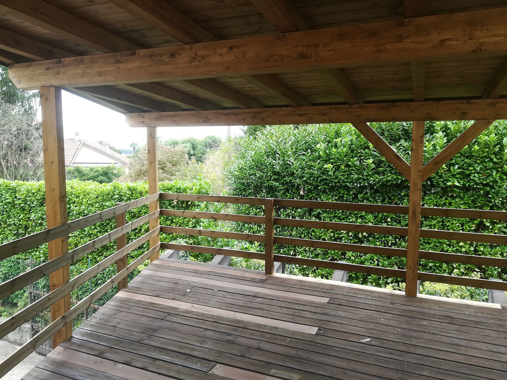 Veranda coperta in legno con ringhiere che si affaccia su arbusti e alberi verdi.