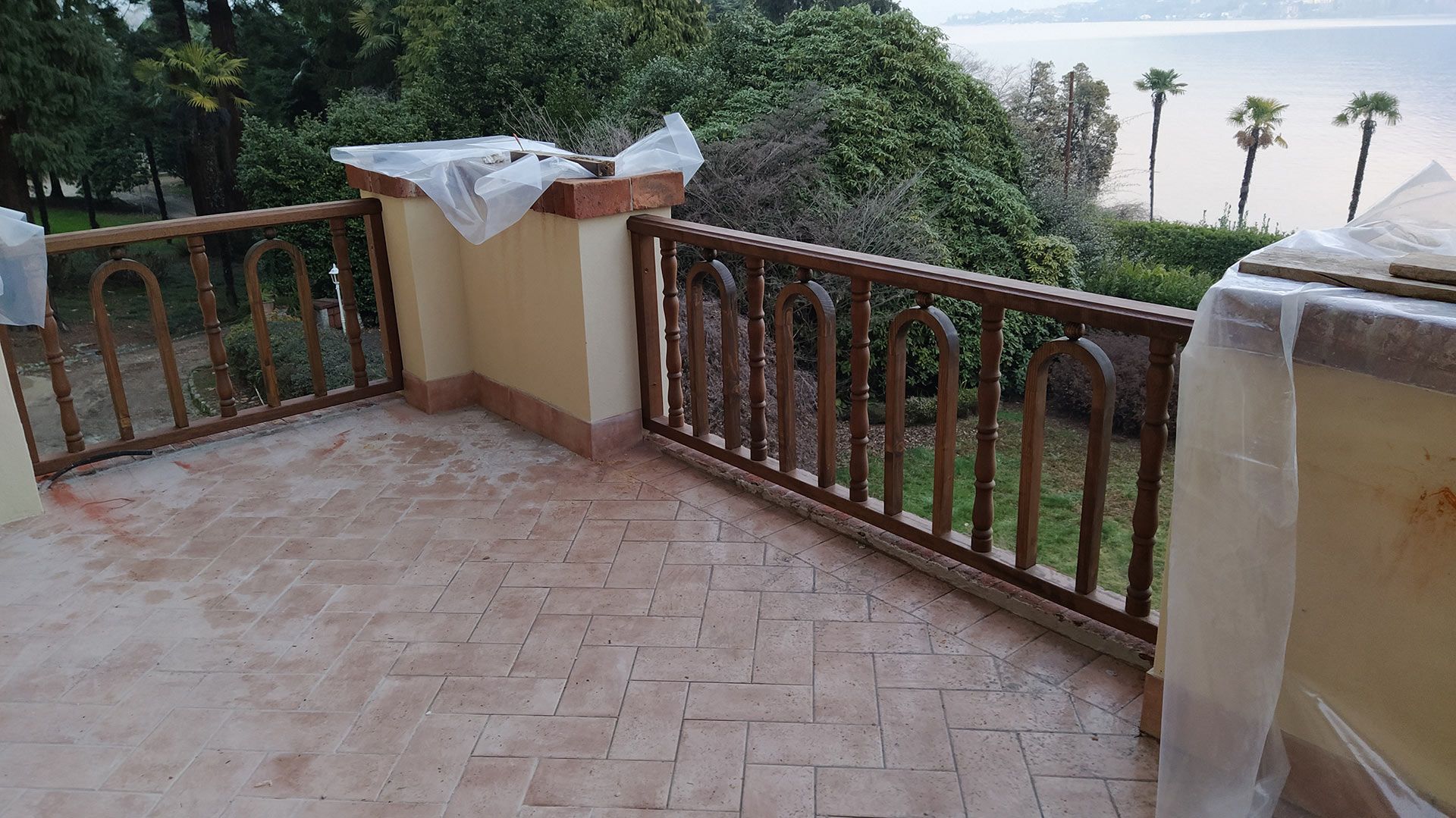 Balcone vuoto piastrellato con ringhiera in legno, affacciato su un giardino verdeggiante e palme al crepuscolo.