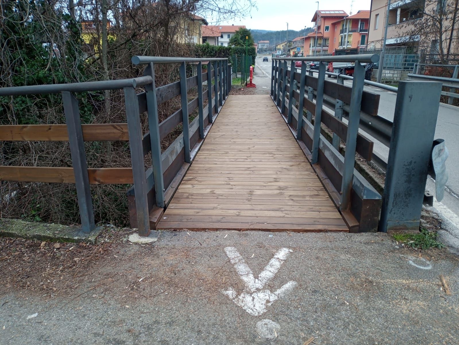 Ponte pedonale in legno con ringhiere in metallo e una freccia bianca dipinta sul marciapiede che vi conduce.