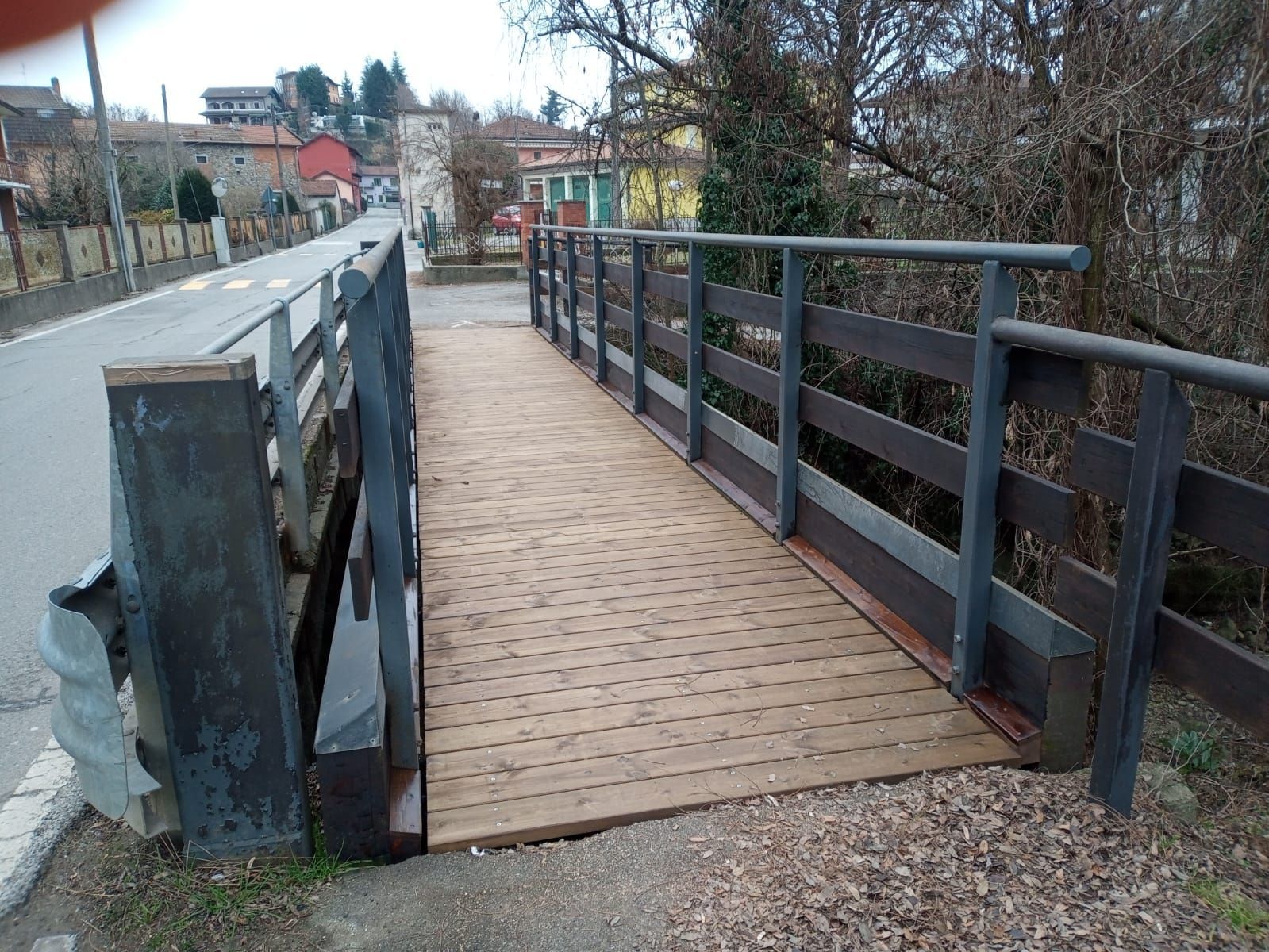 Ponte pedonale in legno con ringhiere metalliche che attraversa un piccolo fiume in un contesto simile a un parco.