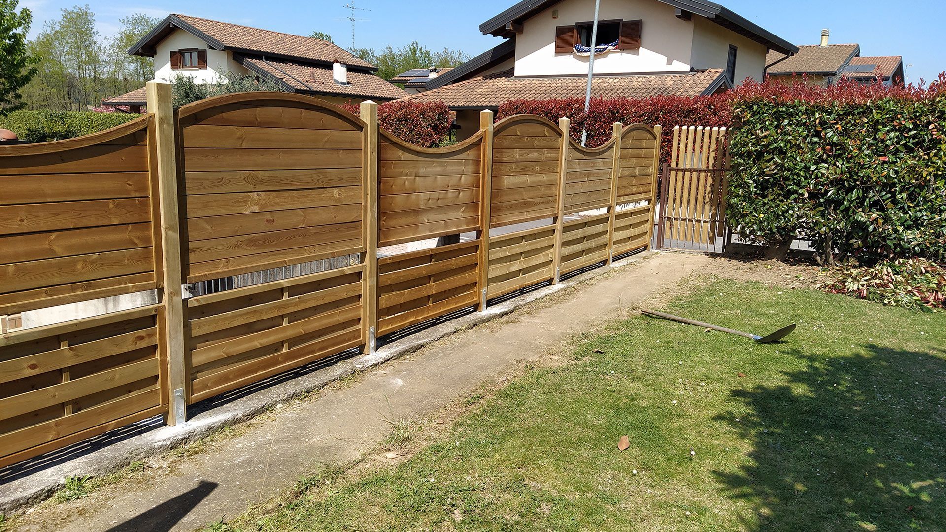 Recinzione da giardino in legno con pannelli curvi, adiacente a un prato e una siepe, con case sullo sfondo.
