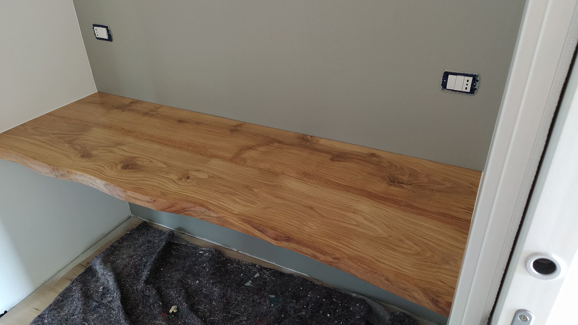 Mensola in legno installata in un piccolo ripostiglio o nicchia con pareti grigie e pavimento in moquette.