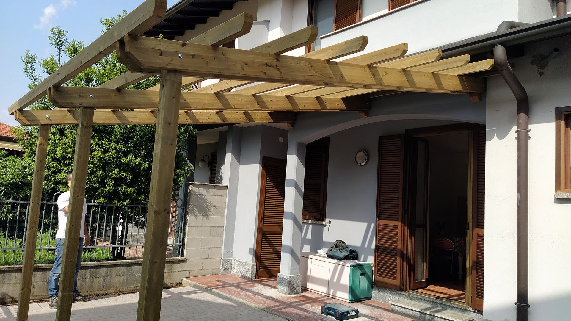 Pergola in legno sopra un patio di casa con porte aperte e piante in vaso esposte al sole