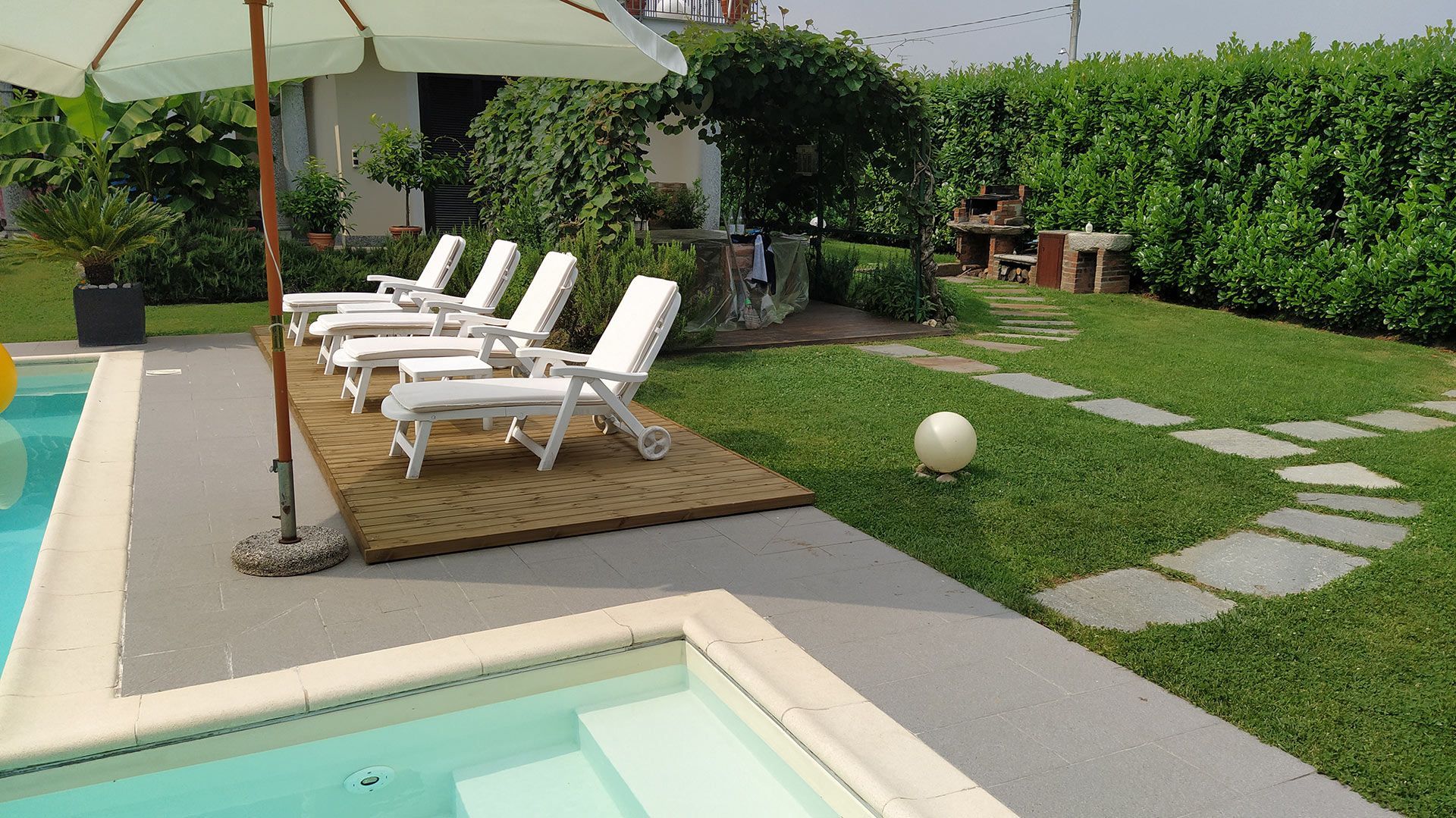 Patio a bordo piscina con lettini bianchi, ombrellone e sentiero in pietra in un verde giardino sul retro.