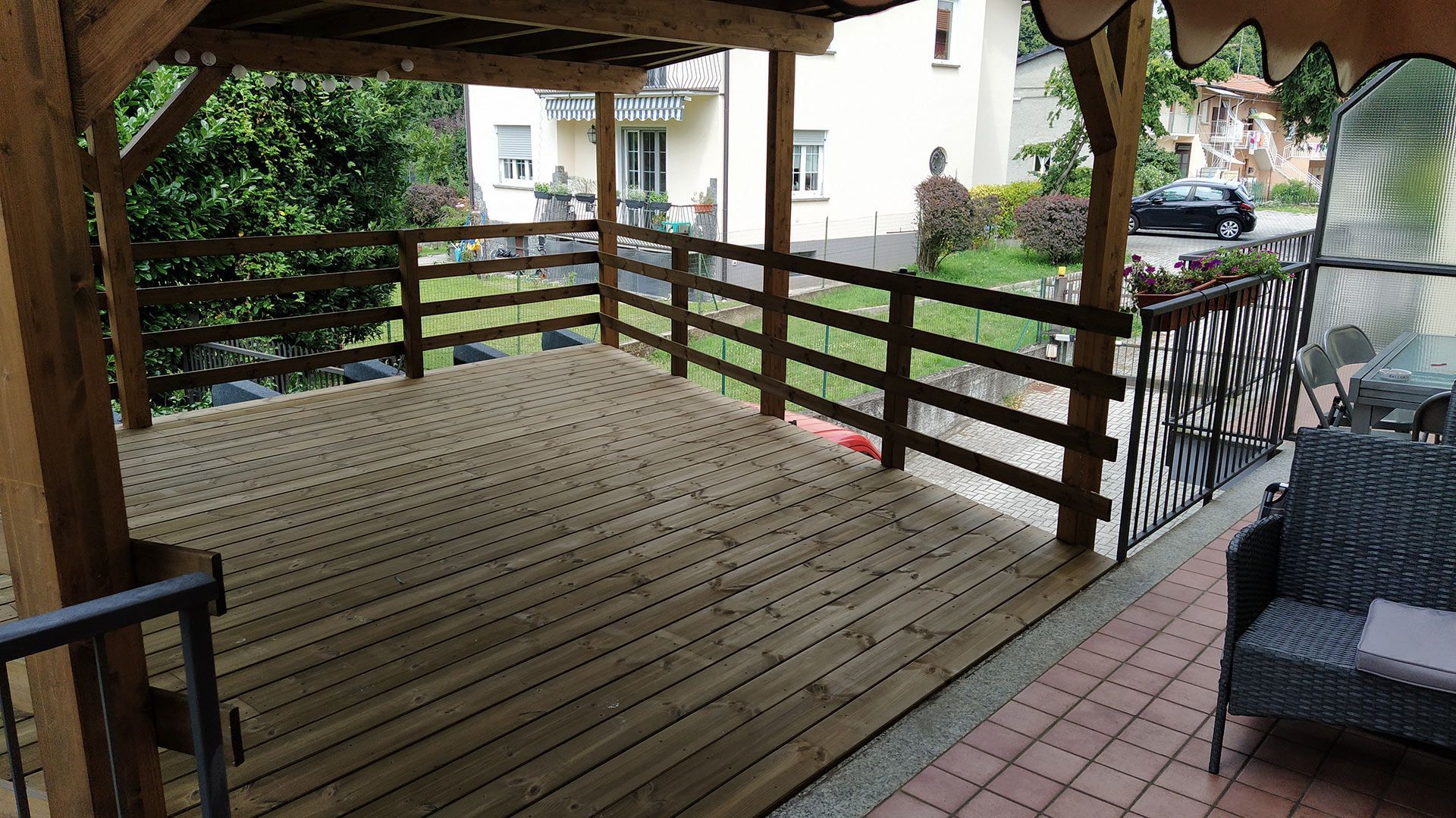 Terrazza in legno coperta con ringhiera accanto a un patio piastrellato, con vista sul verde e su un'auto parcheggiata.
