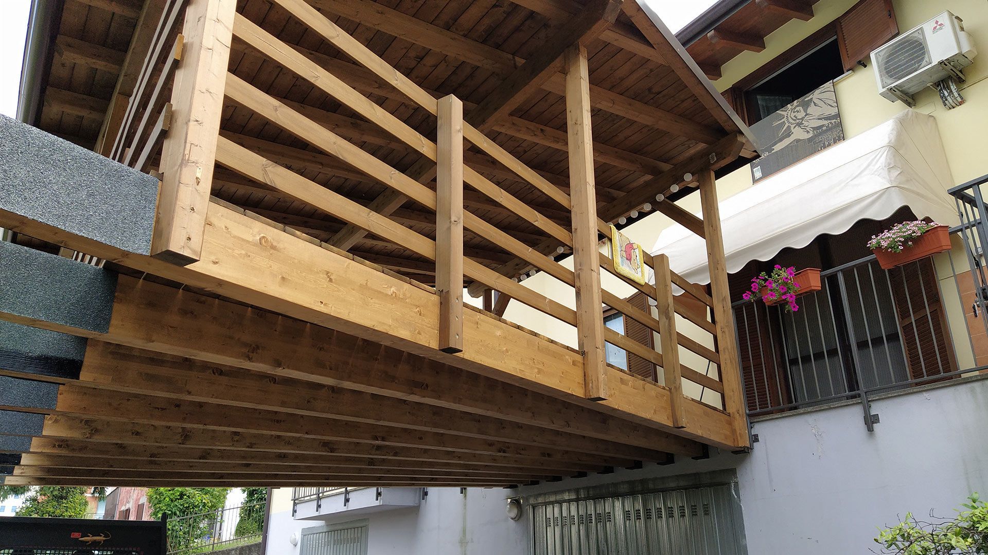 Balcone o terrazza in legno con ringhiera, coperta da una tettoia e annessa a un edificio, adiacente a una scala esterna.