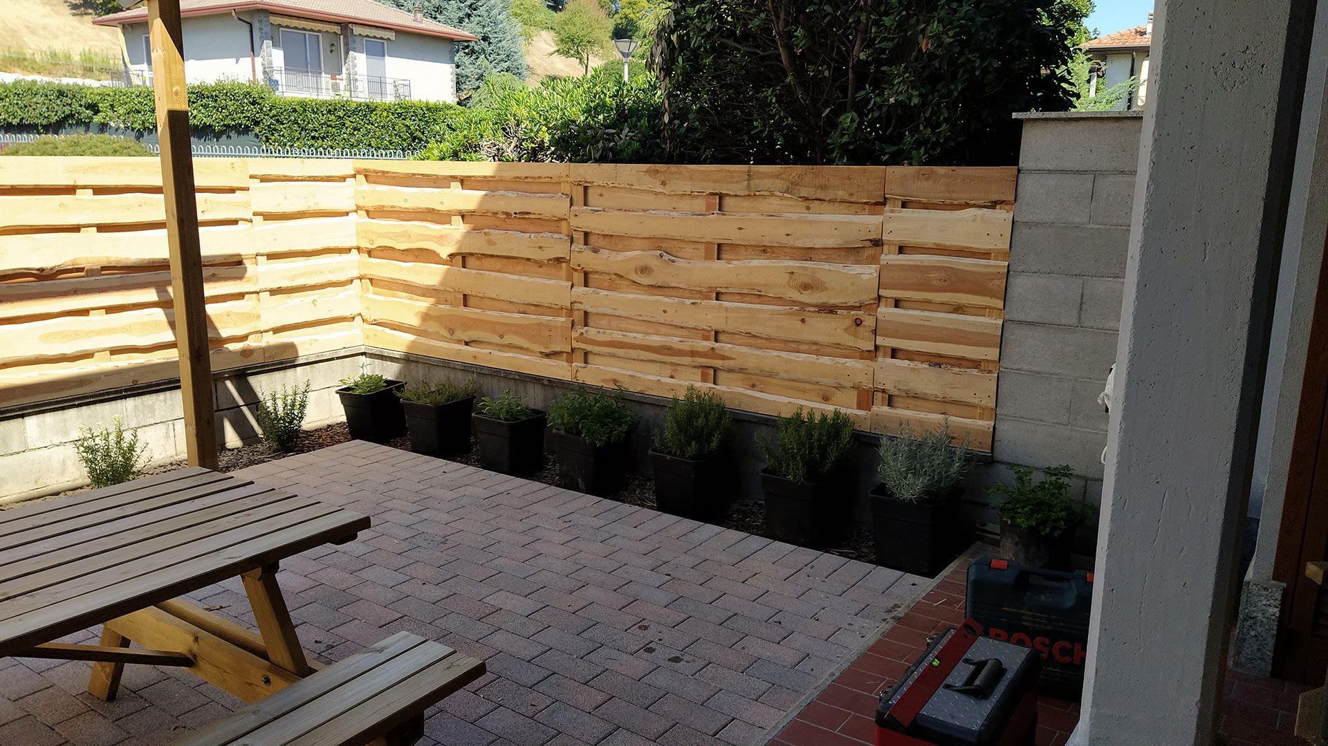 Patio sul retro con recinzione in legno, pavimento pavimentato, tavolo da picnic e piante in vaso esposte al sole.