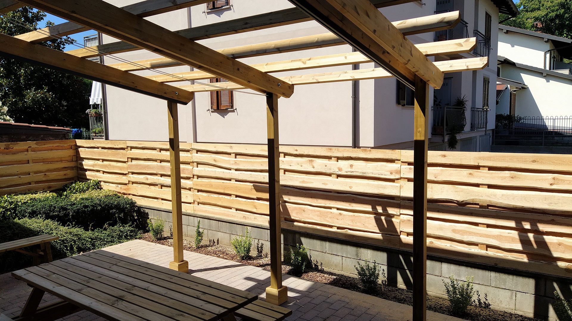 Pergola in legno sopra un patio pavimentato, accanto a una recinzione a doghe che garantisce la privacy e a una parete beige della casa.