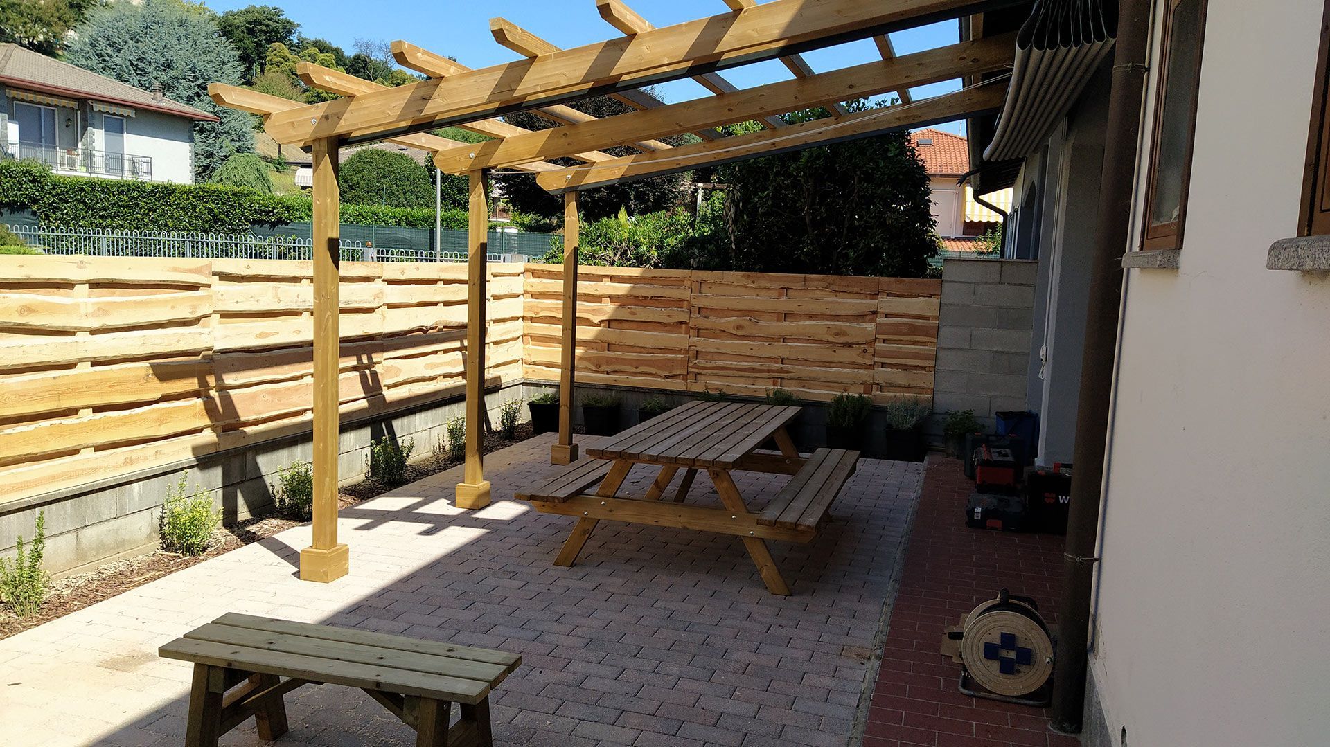 Patio con pergola in legno, tavolo da picnic, piante in vaso e pavimentazione in pietra soleggiata accanto a una casa