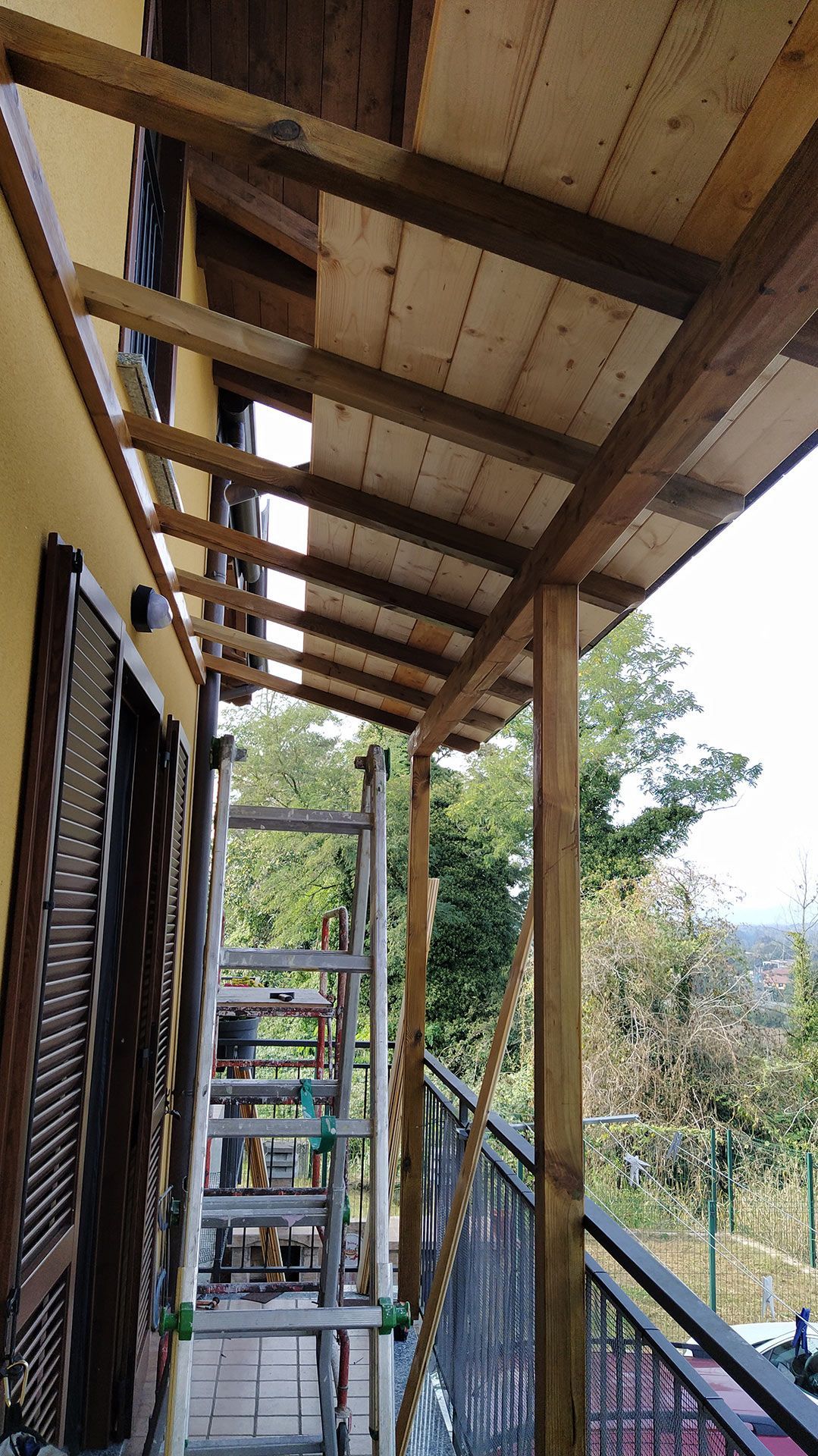 Scala esterna coperta accanto a una terrazza in legno con ringhiere in metallo e alberi sullo sfondo.