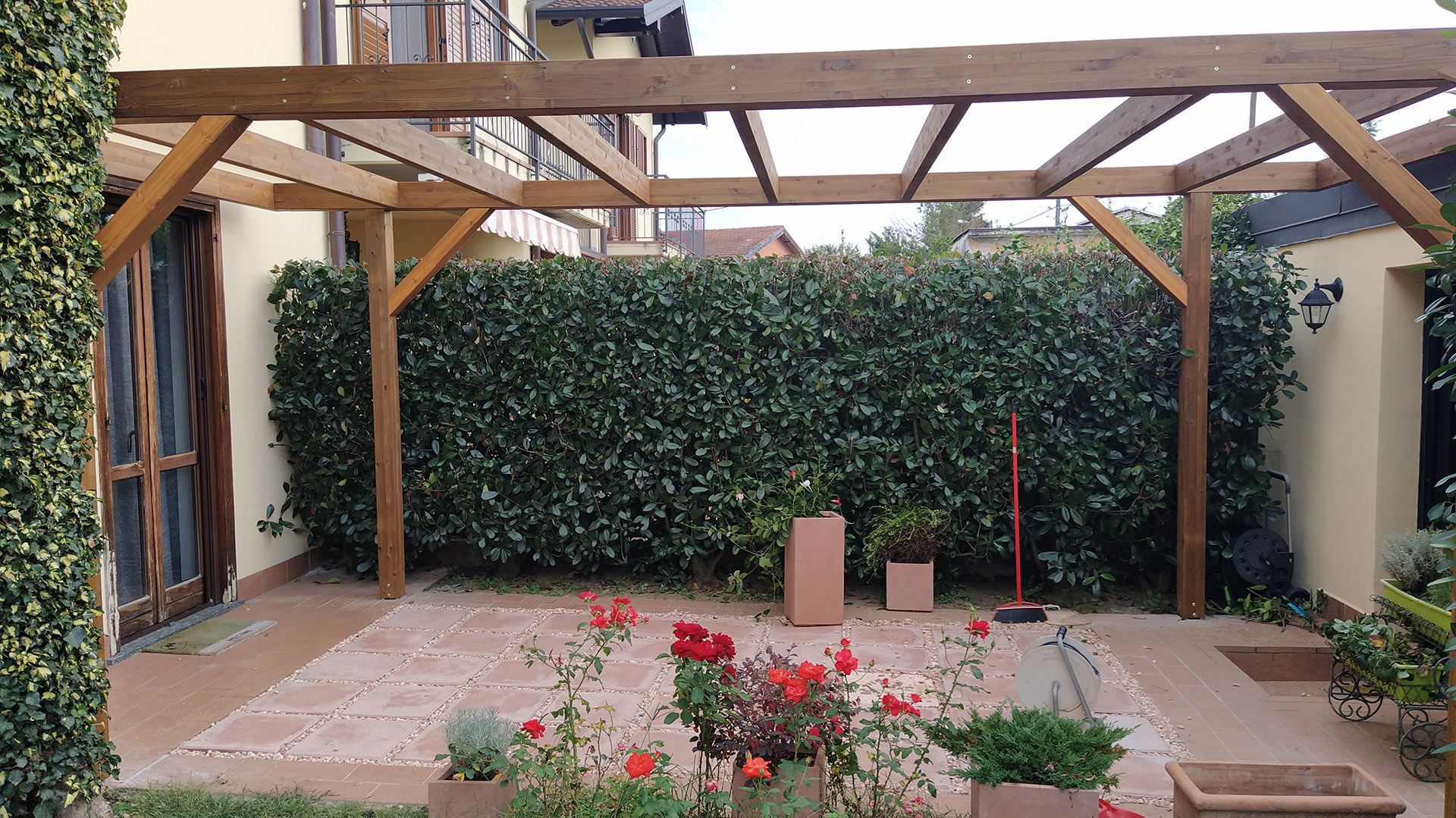 Patio coperto nel cortile sul retro con pergola in legno, parete ricoperta di edera, piante in vaso e fiori rossi a terra.