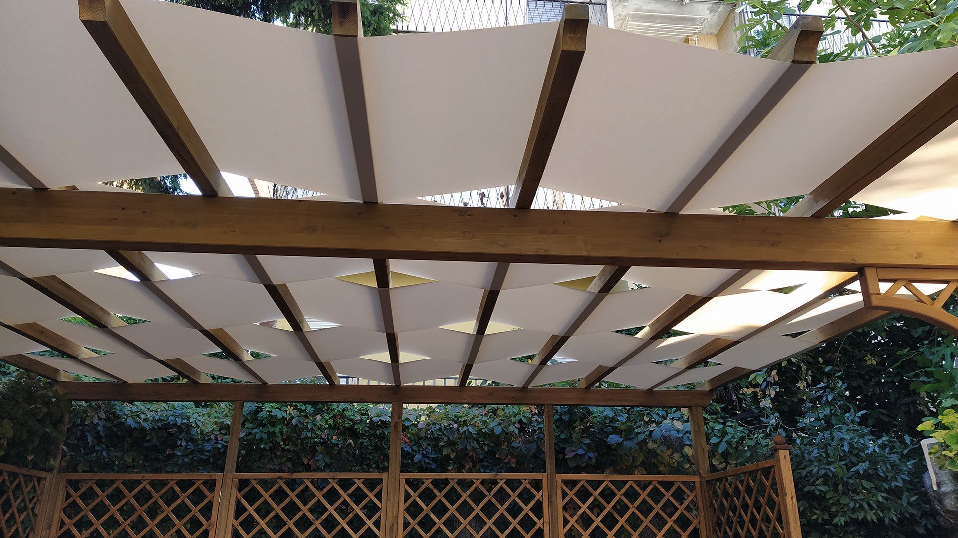Tettoia bianca a pergola con travi scure sopra una recinzione a traliccio in legno e alberi frondosi all'esterno