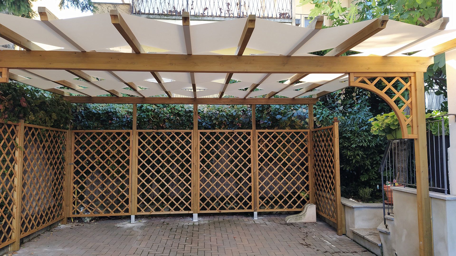 Pergola in legno con traliccio e tetto traslucido in un piccolo cortile pavimentato con vegetazione alle spalle.