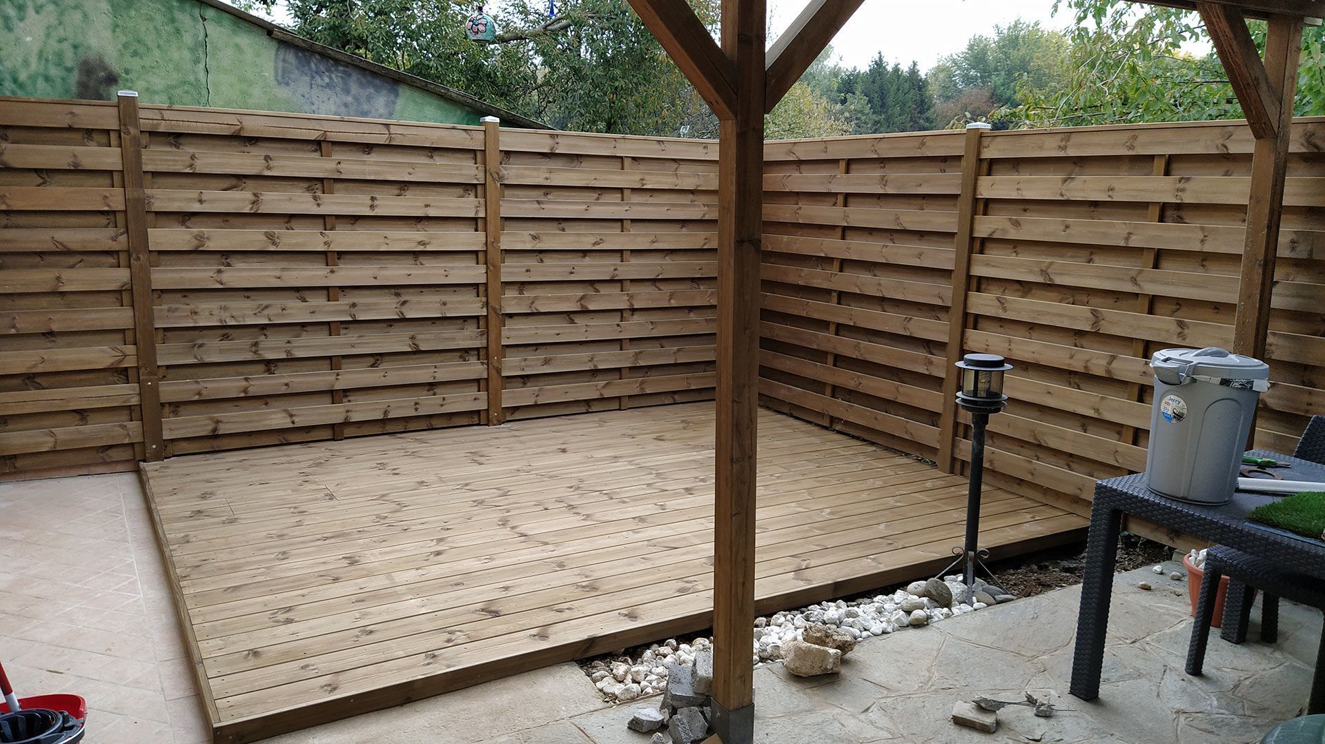 Terrazza in legno sul retro della casa con recinzione per la privacy, palo di sostegno del tetto e piccola area patio sulla destra.